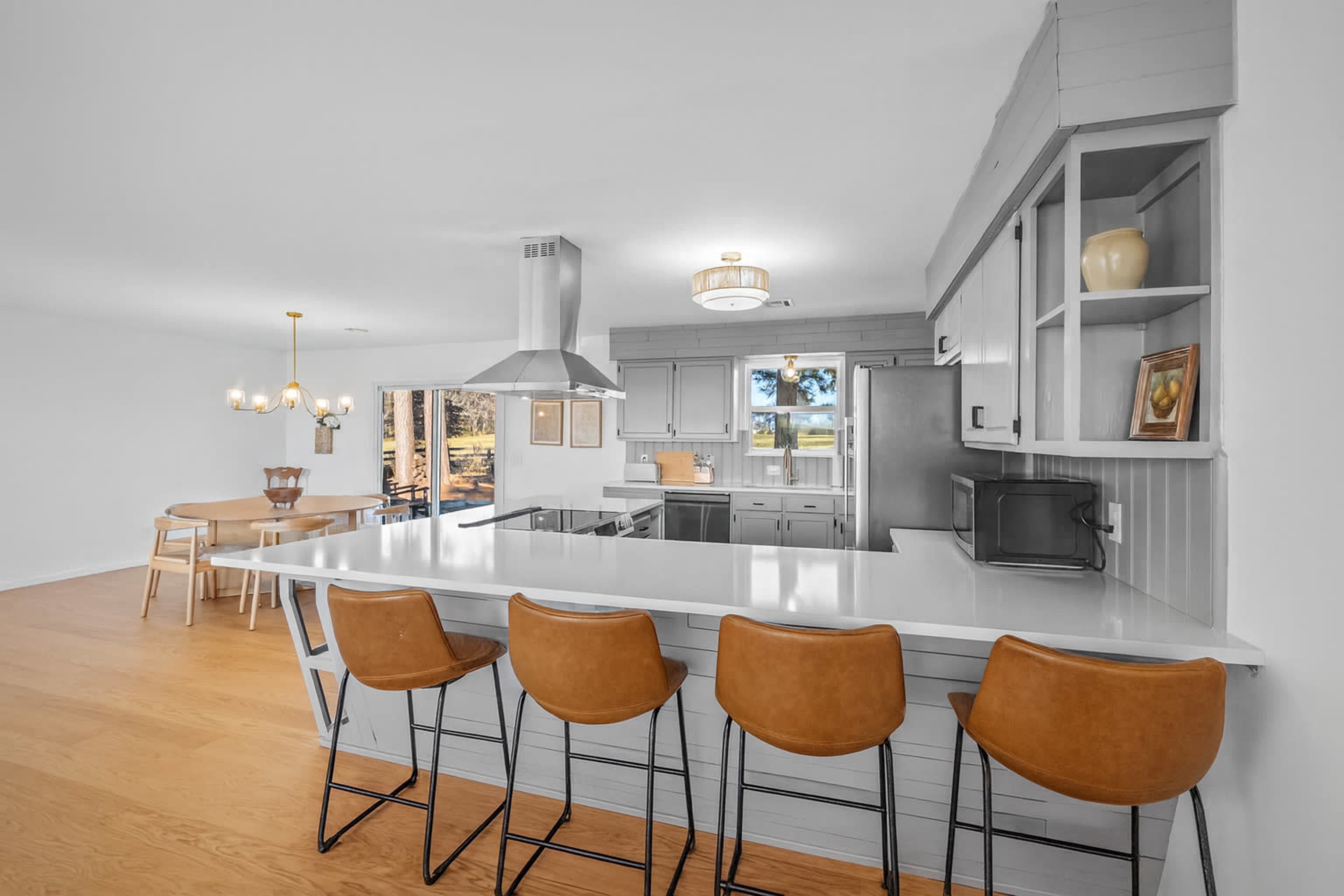 A modern kitchen features a large island with four bar stools, stainless steel appliances, and a dining area in the background with a wooden table and chairs.