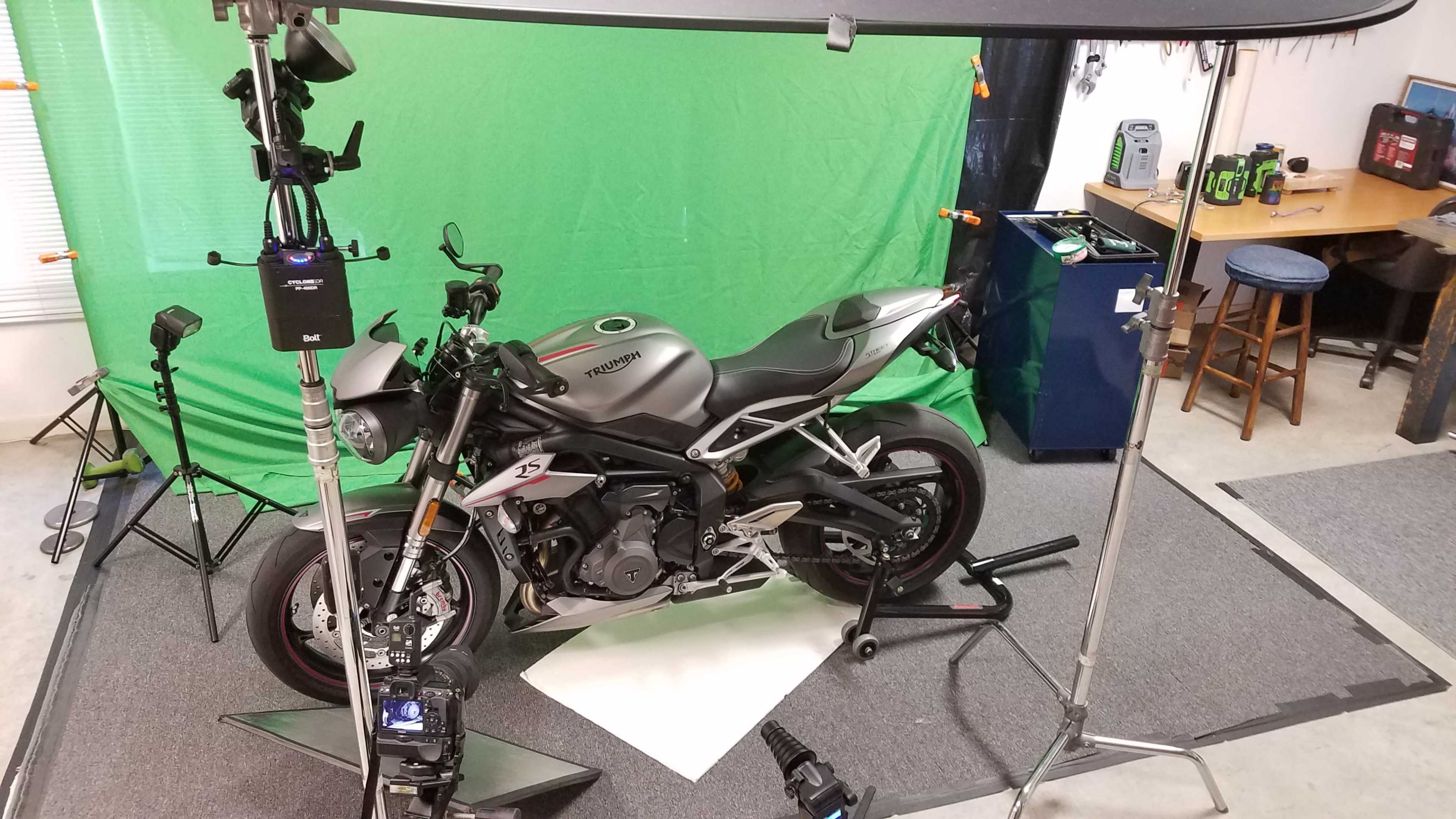 A motorcycle is positioned on a stand in front of a green screen, surrounded by photography equipment in a workshop.