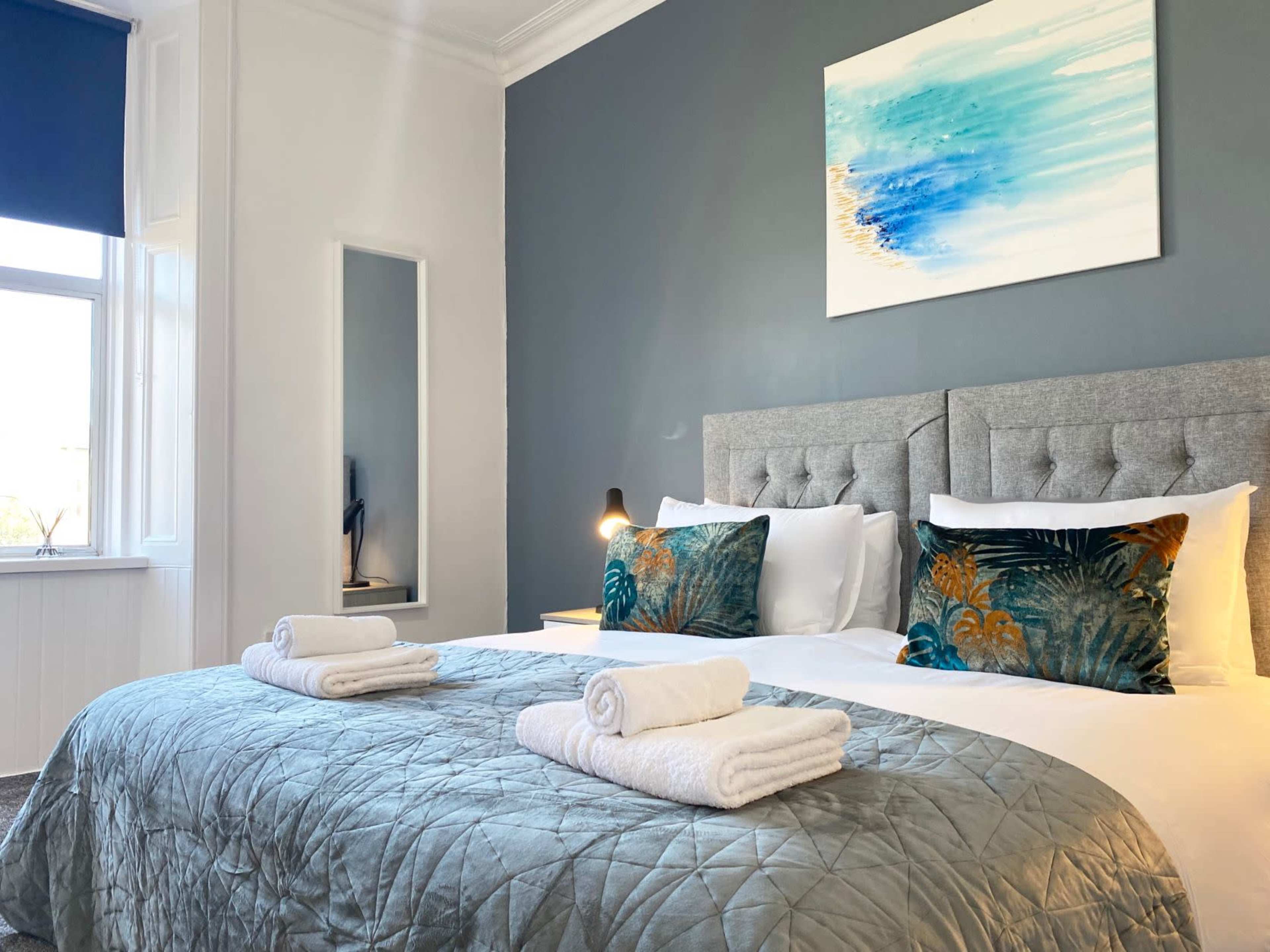 A stylish hotel room with two beds, each adorned with white towels, a decorative throw blanket, and a piece of abstract wall art above a gray accent wall.