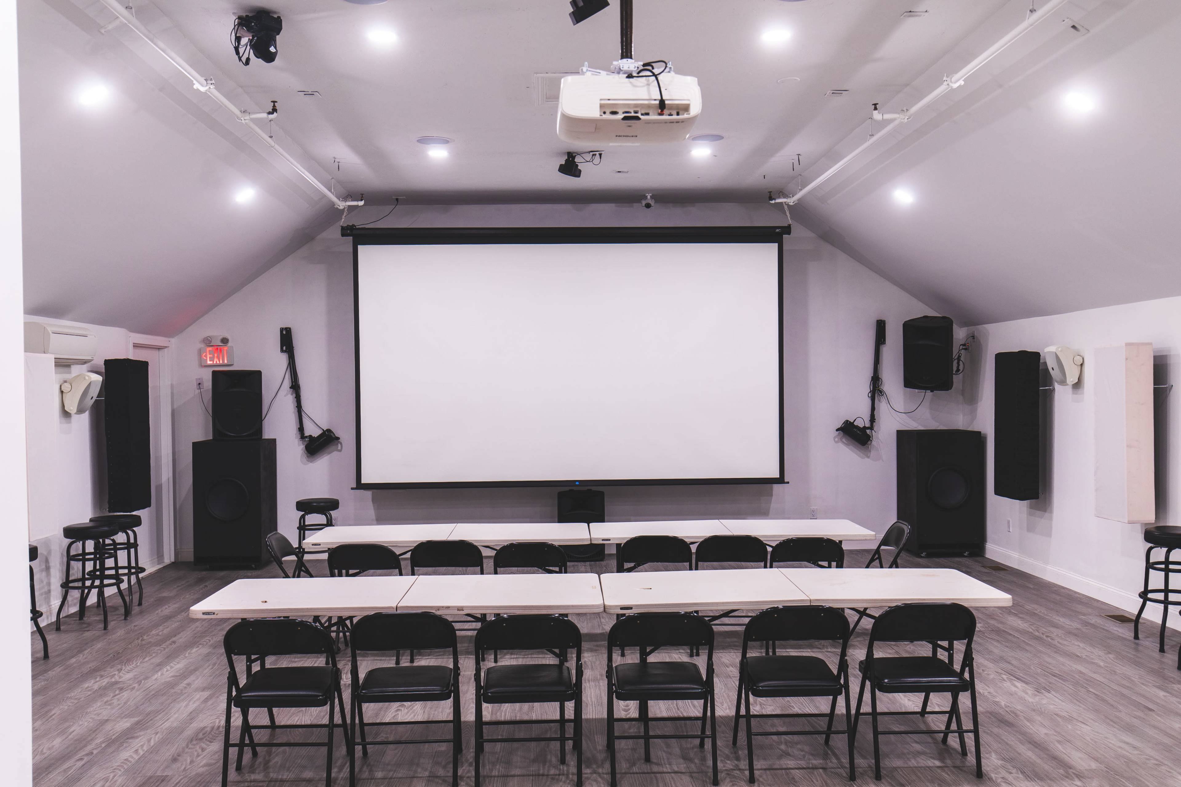 A spacious room is equipped with a large projection screen, rows of chairs, and audio speakers on either side.