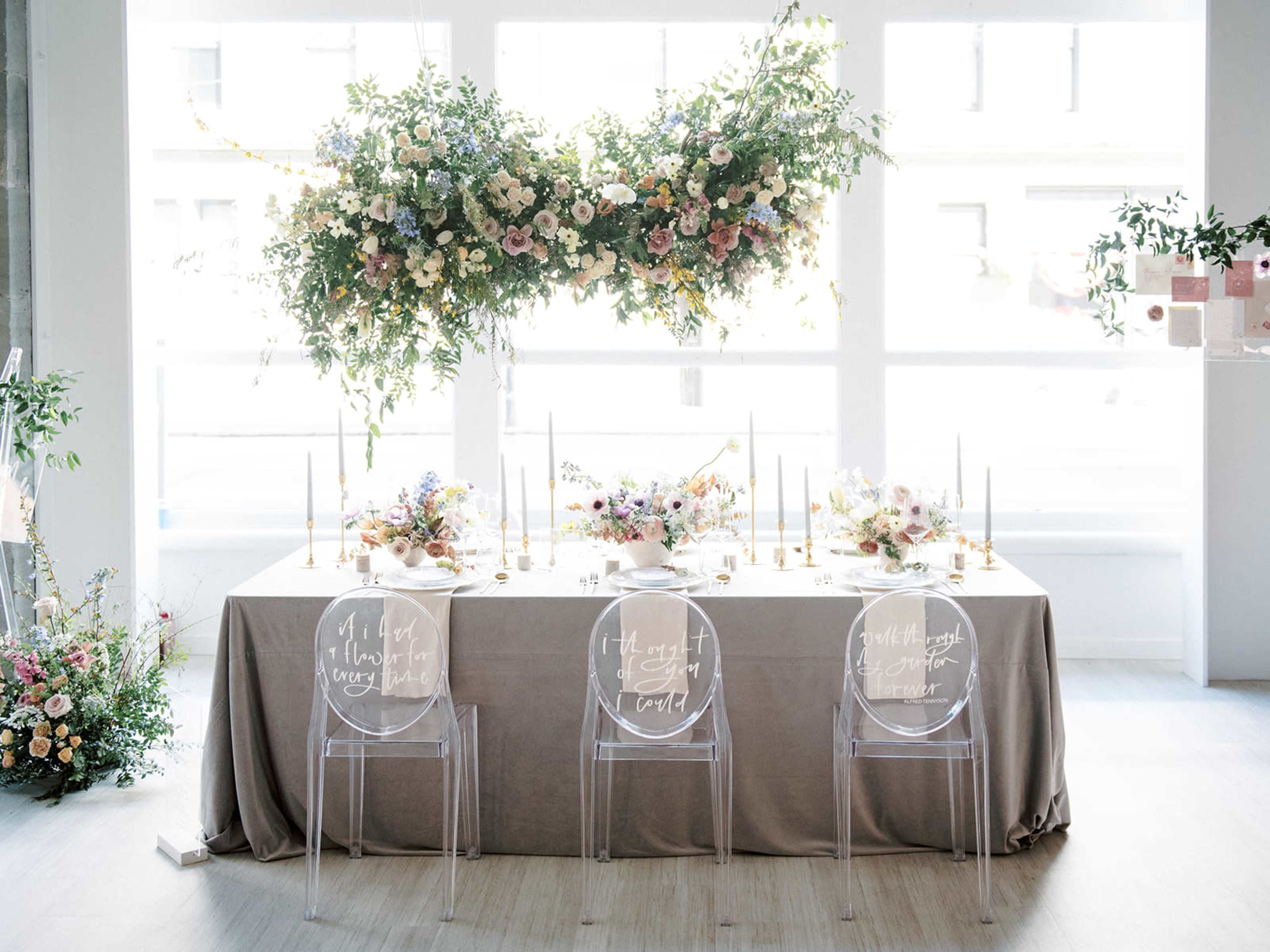 A styled banquet table is set with floral arrangements and candles, flanked by three transparent chairs with handwritten text.