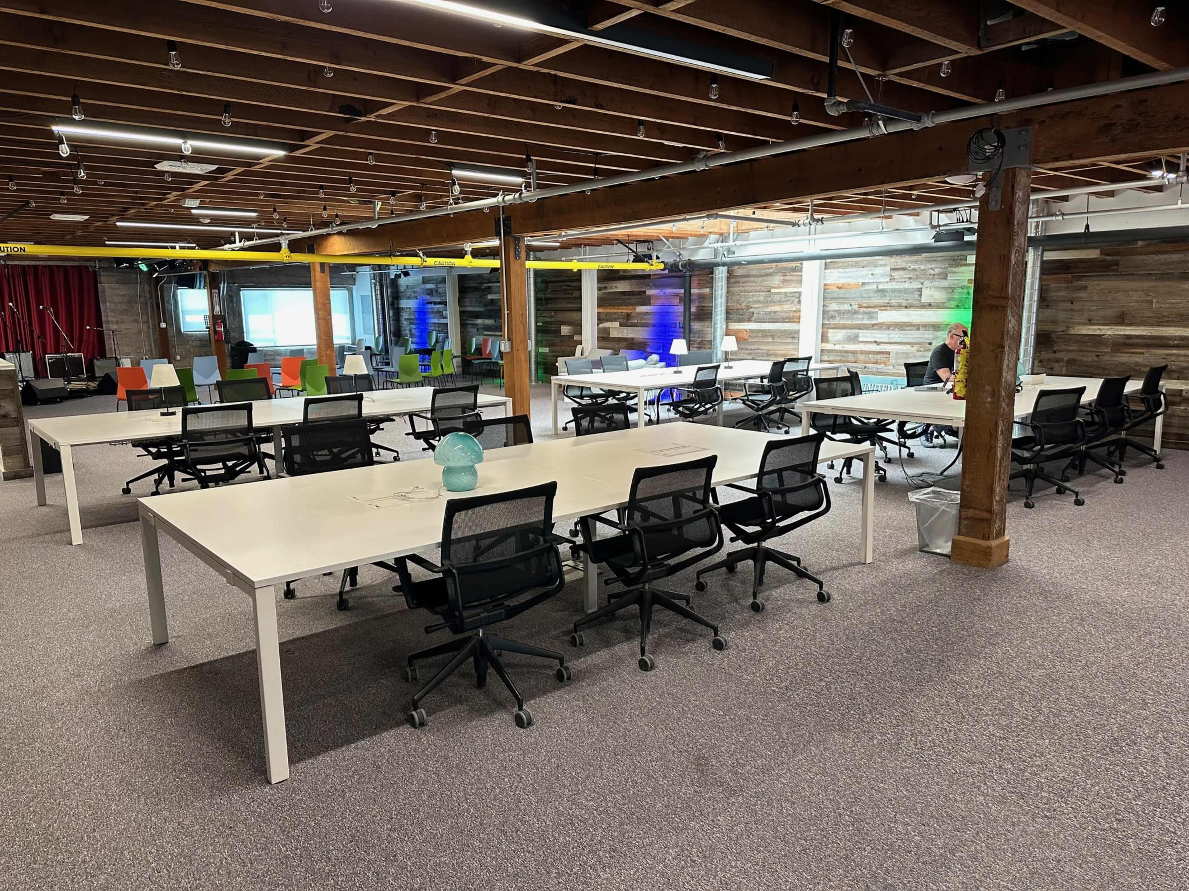 A spacious, modern office area features multiple white tables with black chairs and colorful seating in the background, illuminated by bright lighting.