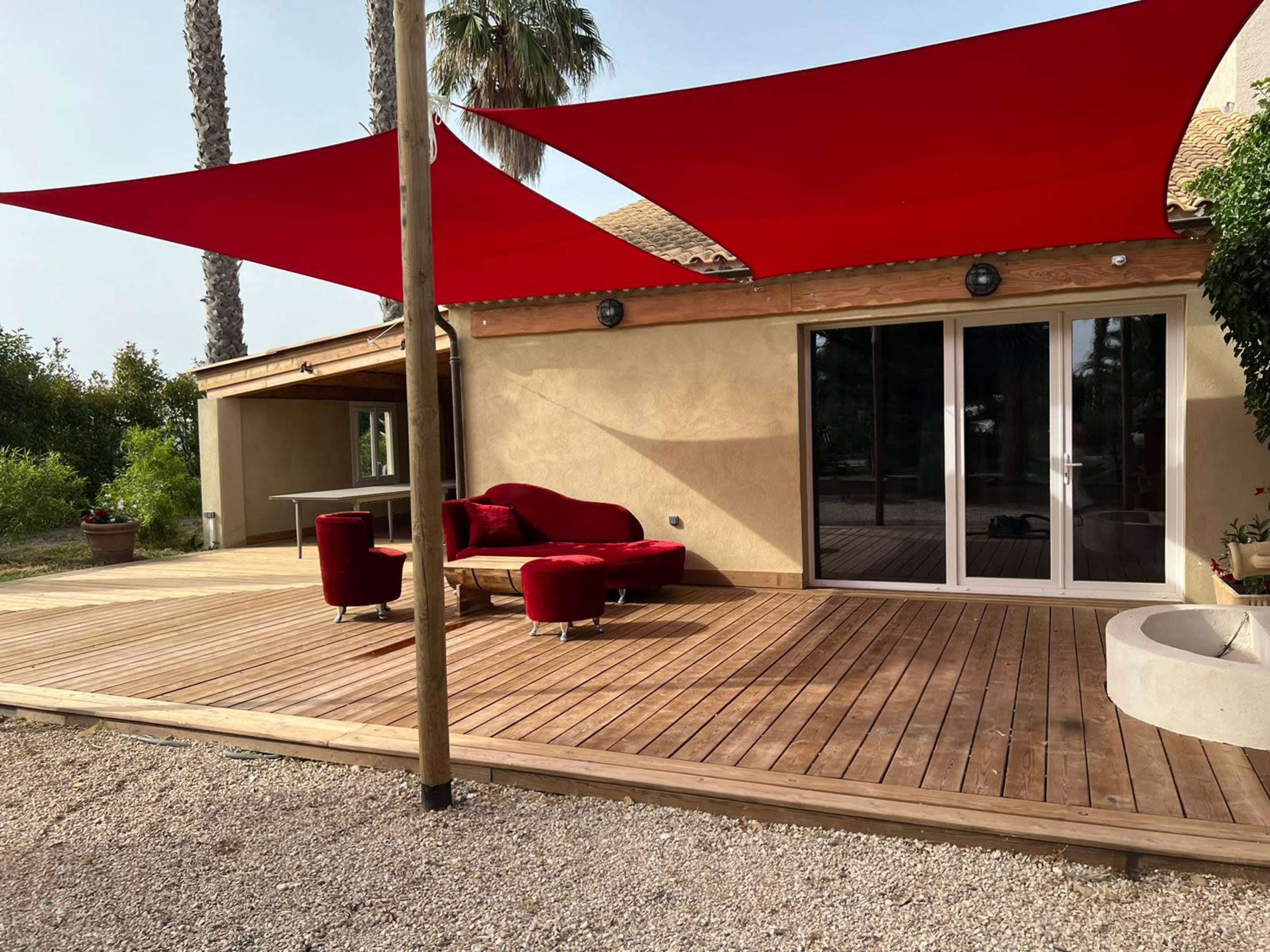 The image shows a wooden deck with red shade sails above, adjacent to a house with large glass doors.