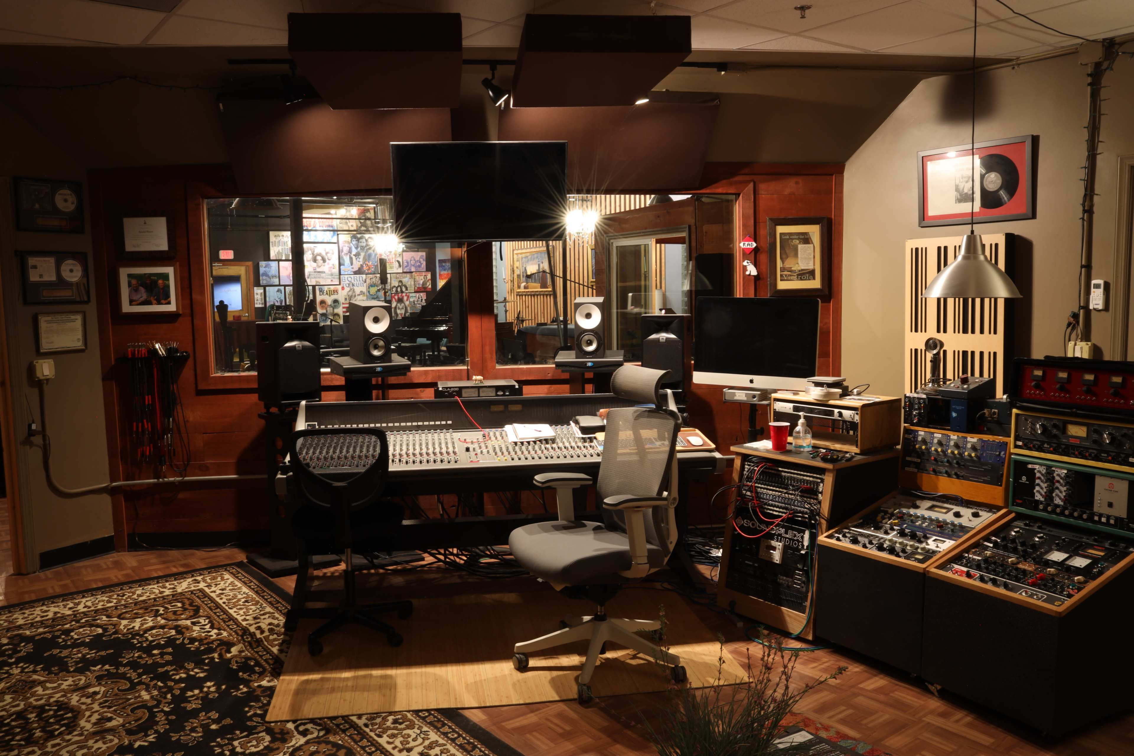 A recording studio with a central mixing desk, two speakers, computer equipment, and various sound processing devices arranged around a plush area rug.