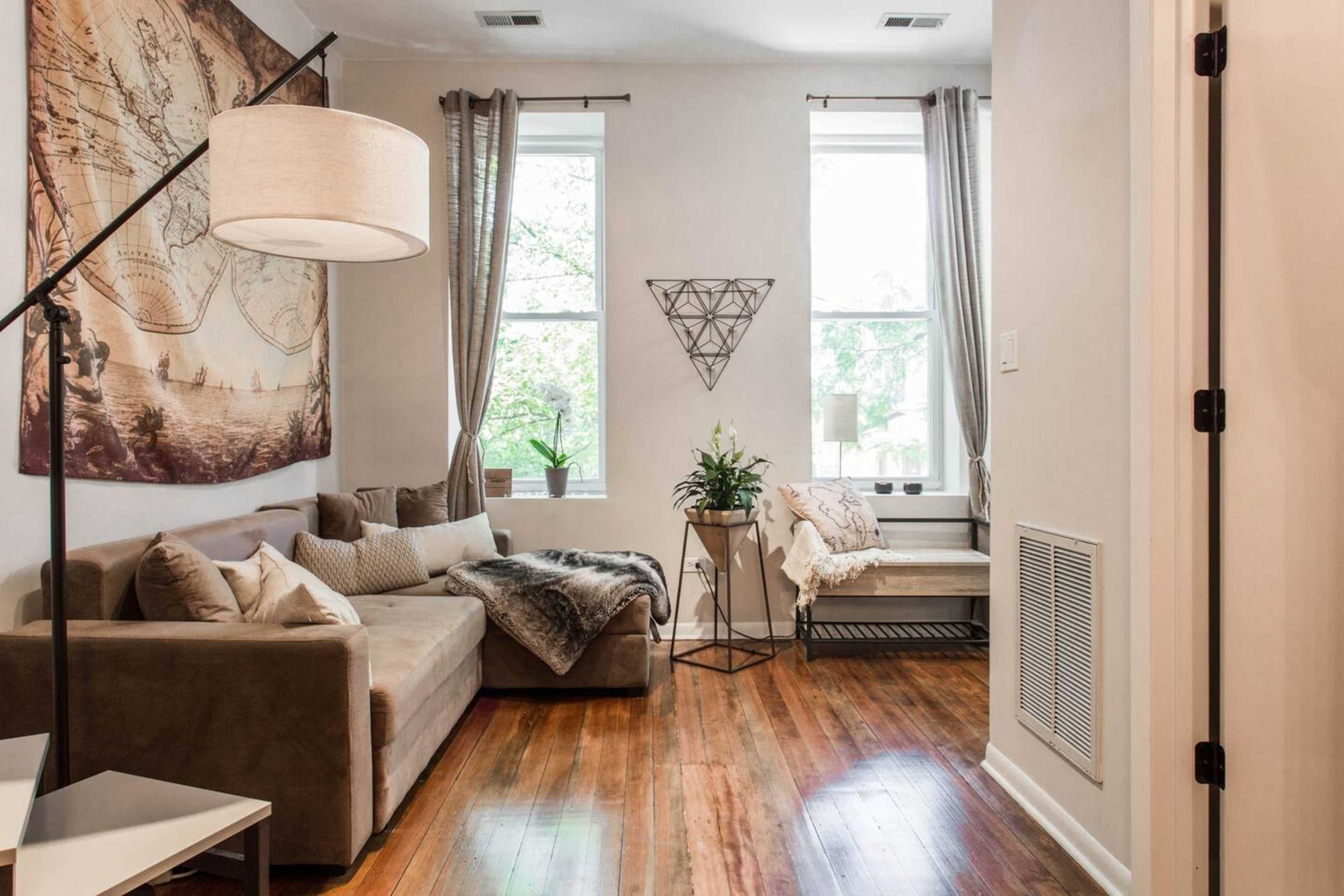 The room features a sofa with cushions, a light fixture, a plant in a corner, and large windows with curtains.