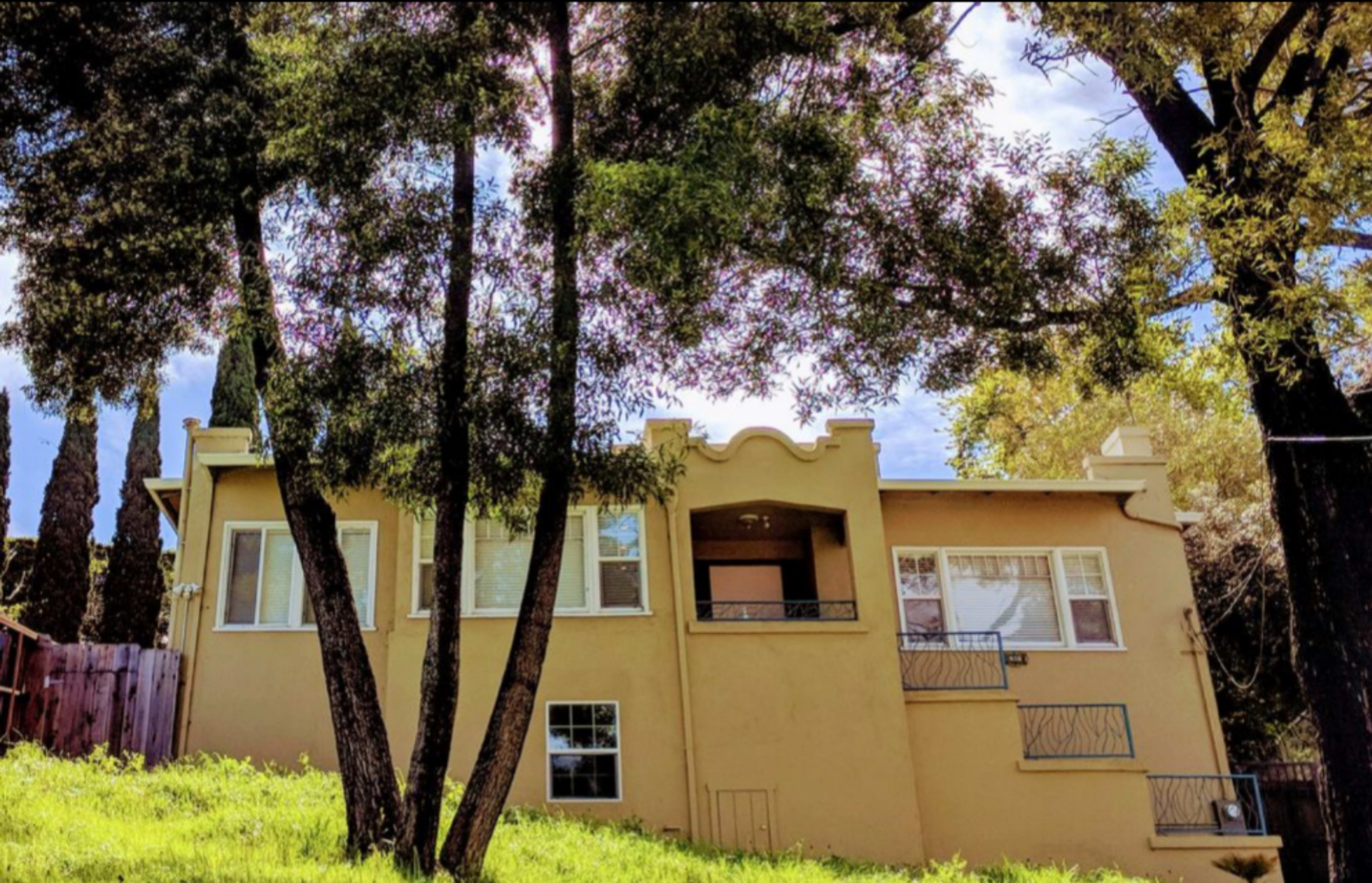 A tan house with large windows and decorative rooflines sits on a grassy slope, surrounded by tall trees.