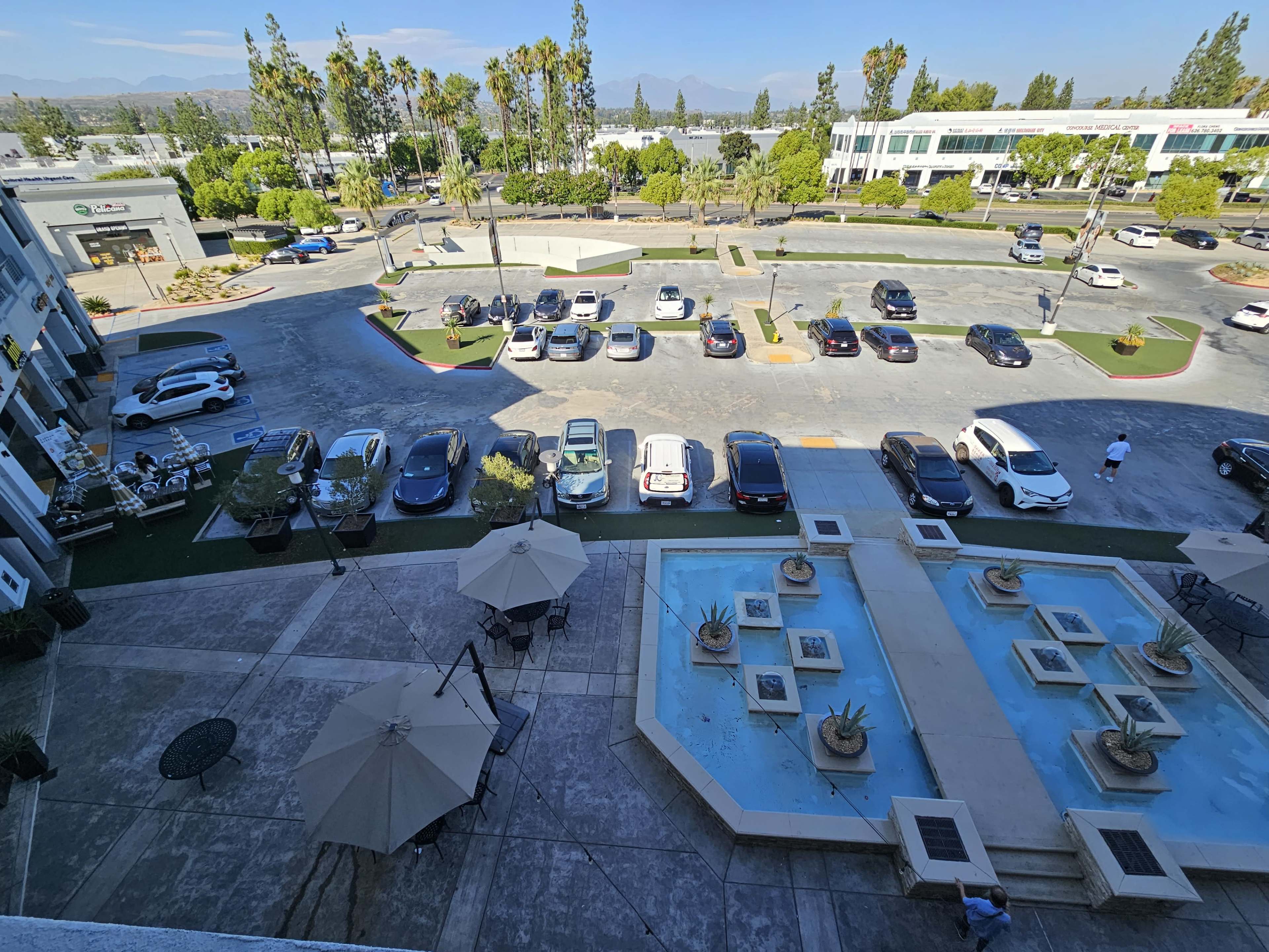 A commercial area with parked cars, landscaped surroundings, and a fountain in the foreground.