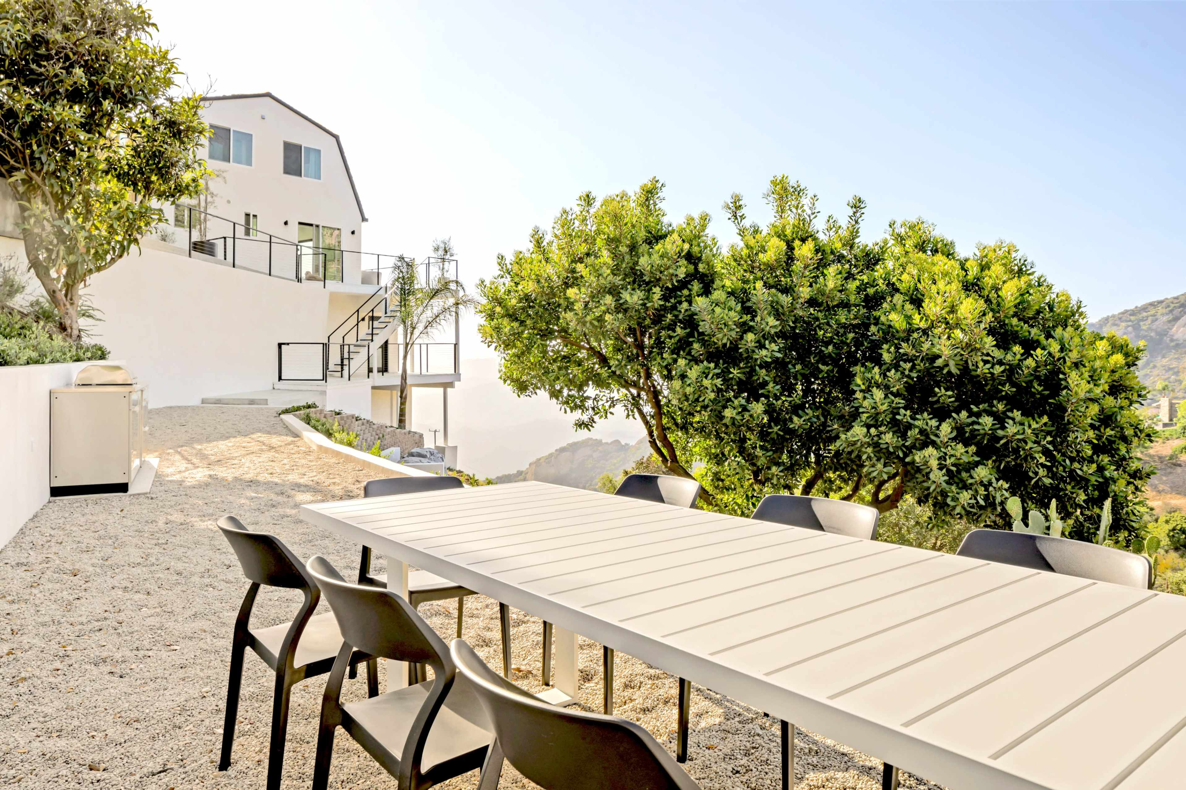 A long, white table with black chairs is set on a gravel patio overlooking a hillside house and lush greenery.