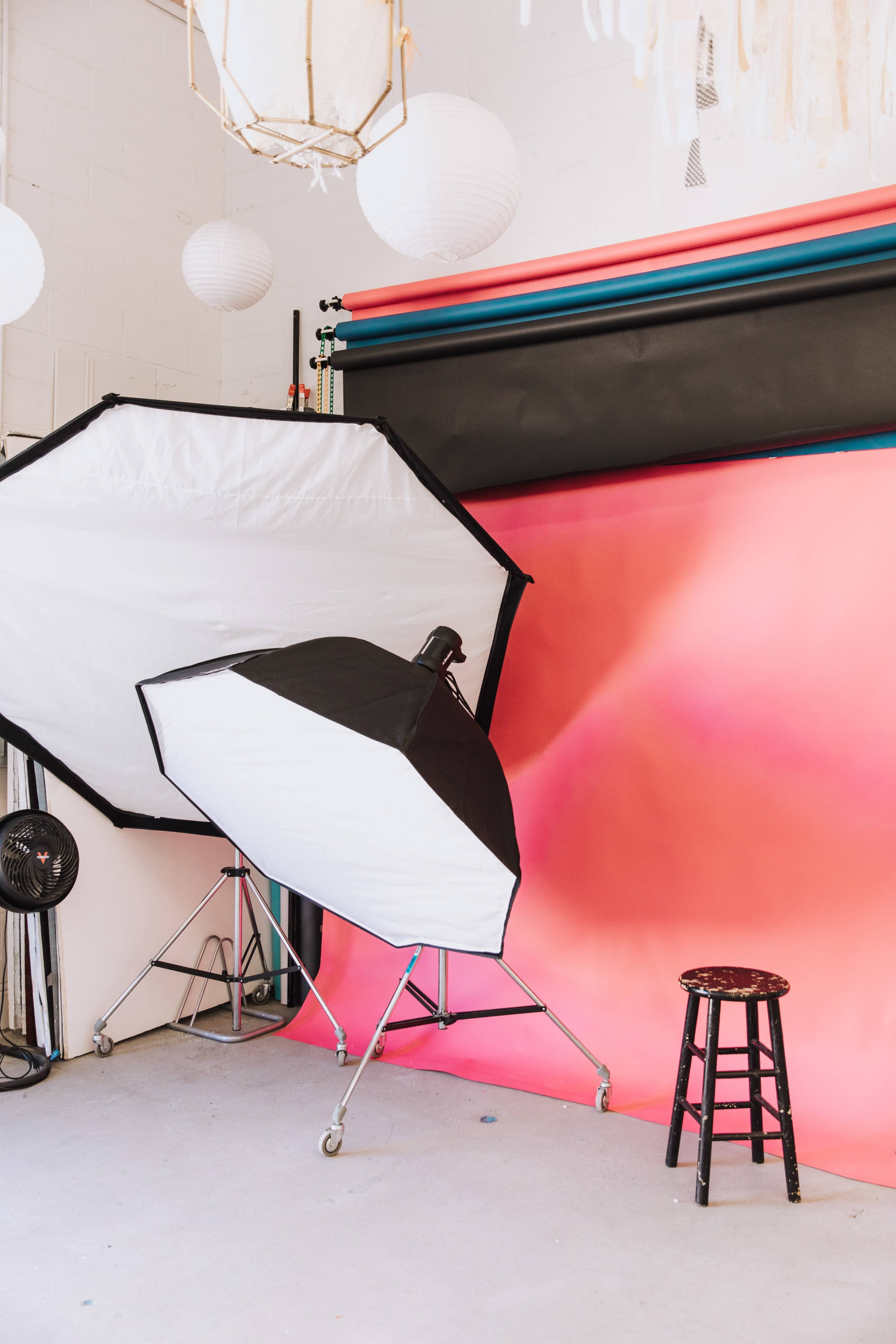 A photography studio features a large softbox lighting setup next to a dark pink and black backdrop, with a wooden stool positioned in front.