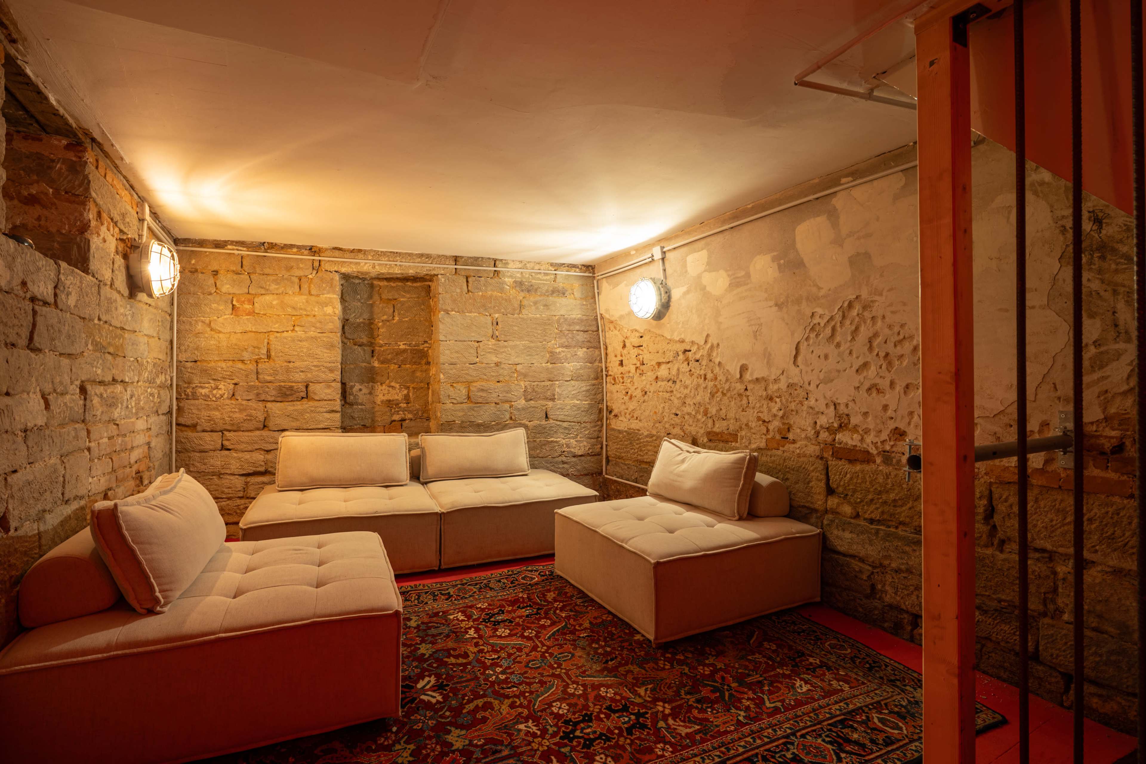 The image shows a secluded basement room with stone walls, furnished with beige couches and a patterned rug.