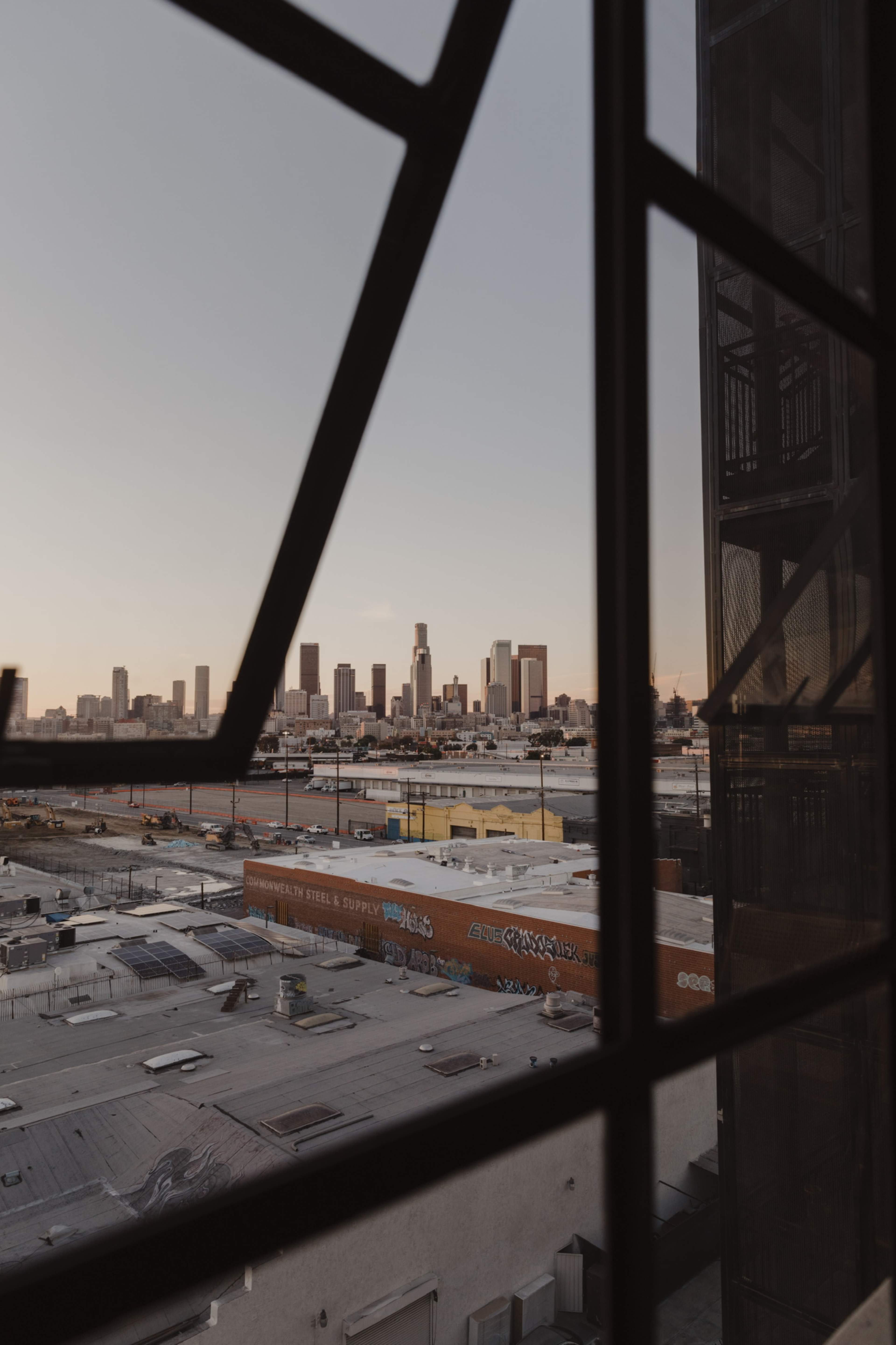 A view of the Los Angeles skyline through a window with a grid pattern, overlooking industrial rooftops and graffiti-covered buildings.