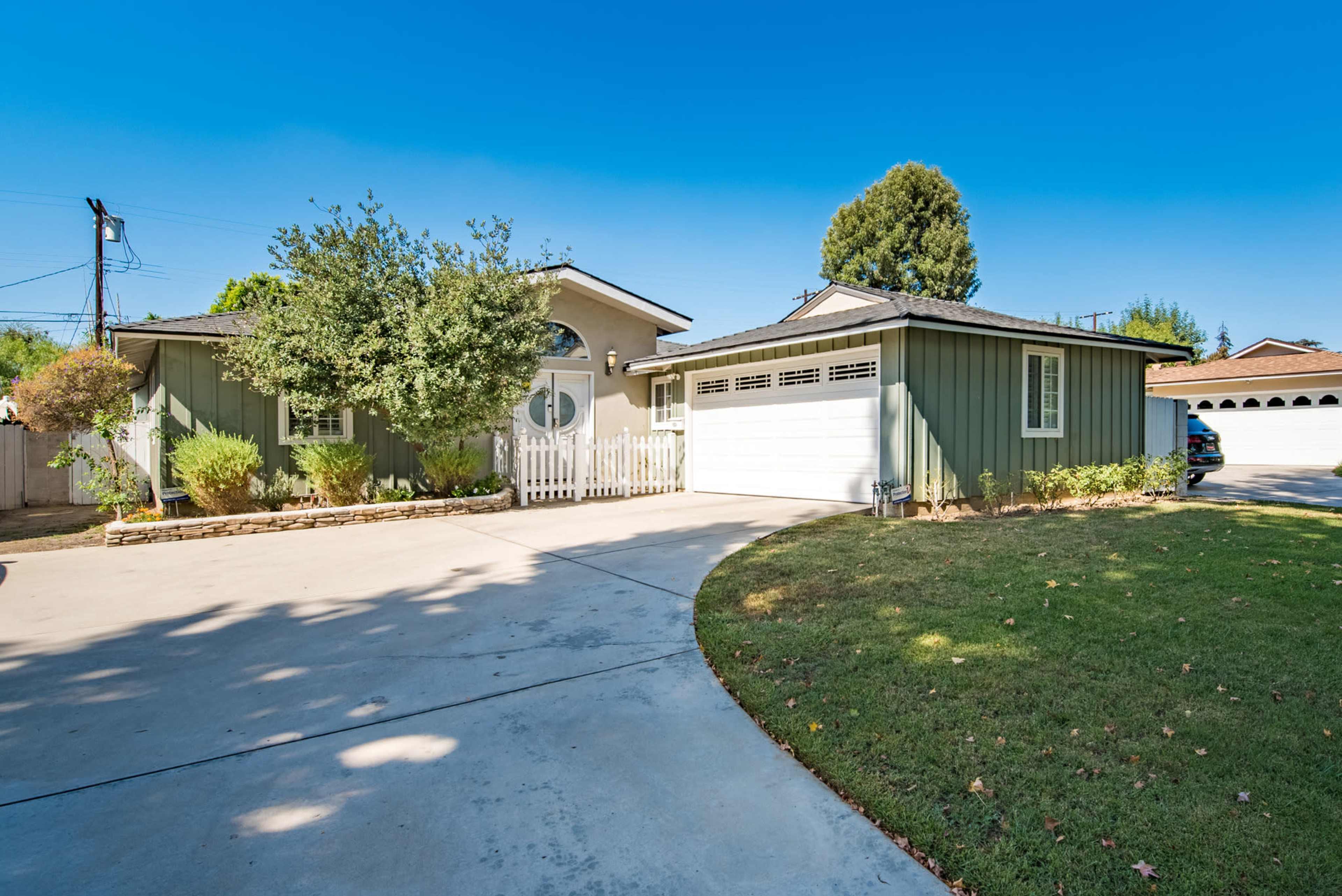 A single-story green house with a white picket fence and a spacious driveway is set against a clear blue sky.