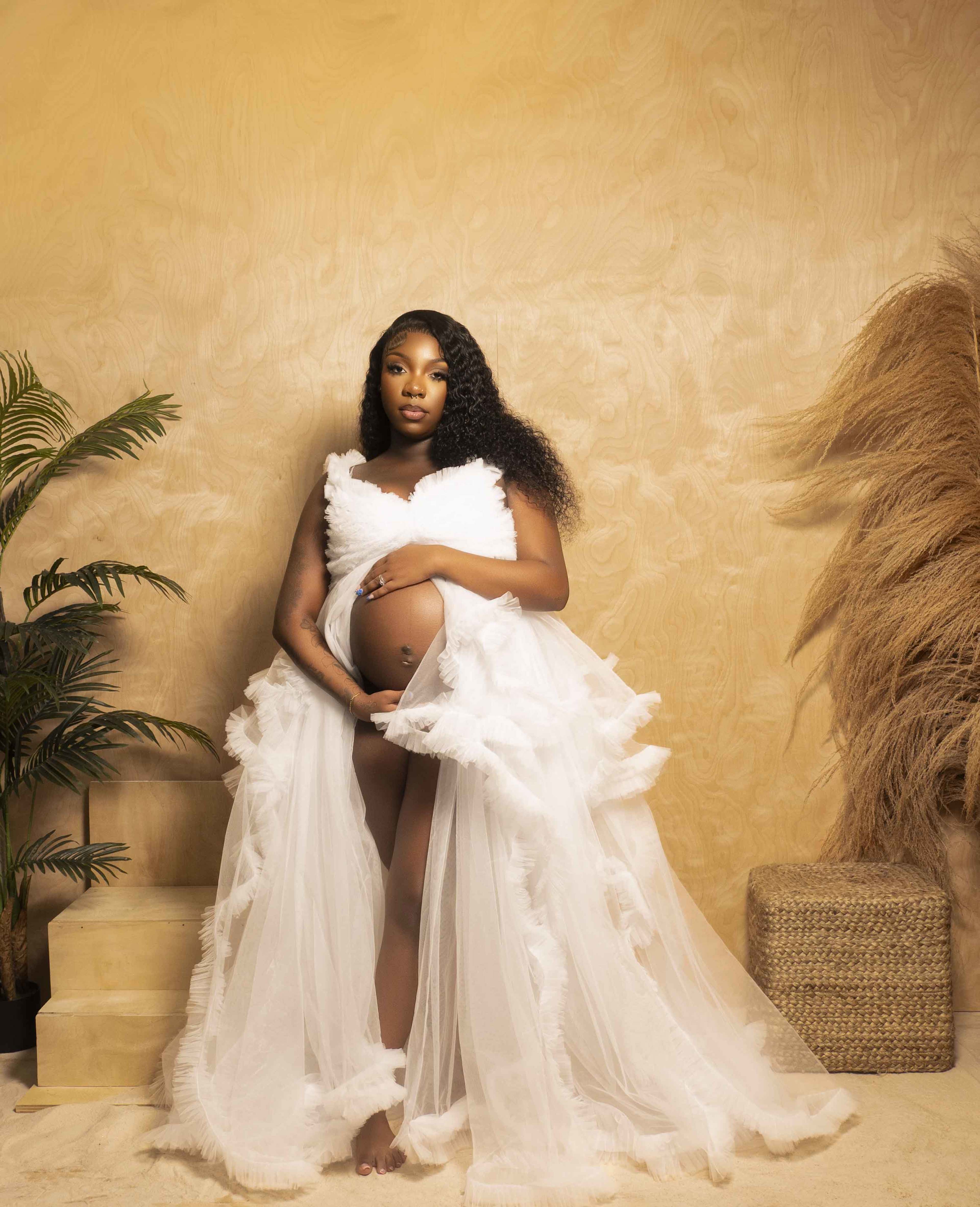 A pregnant woman in a flowing white dress poses against a textured beige backdrop with decorative plants and textured elements nearby.