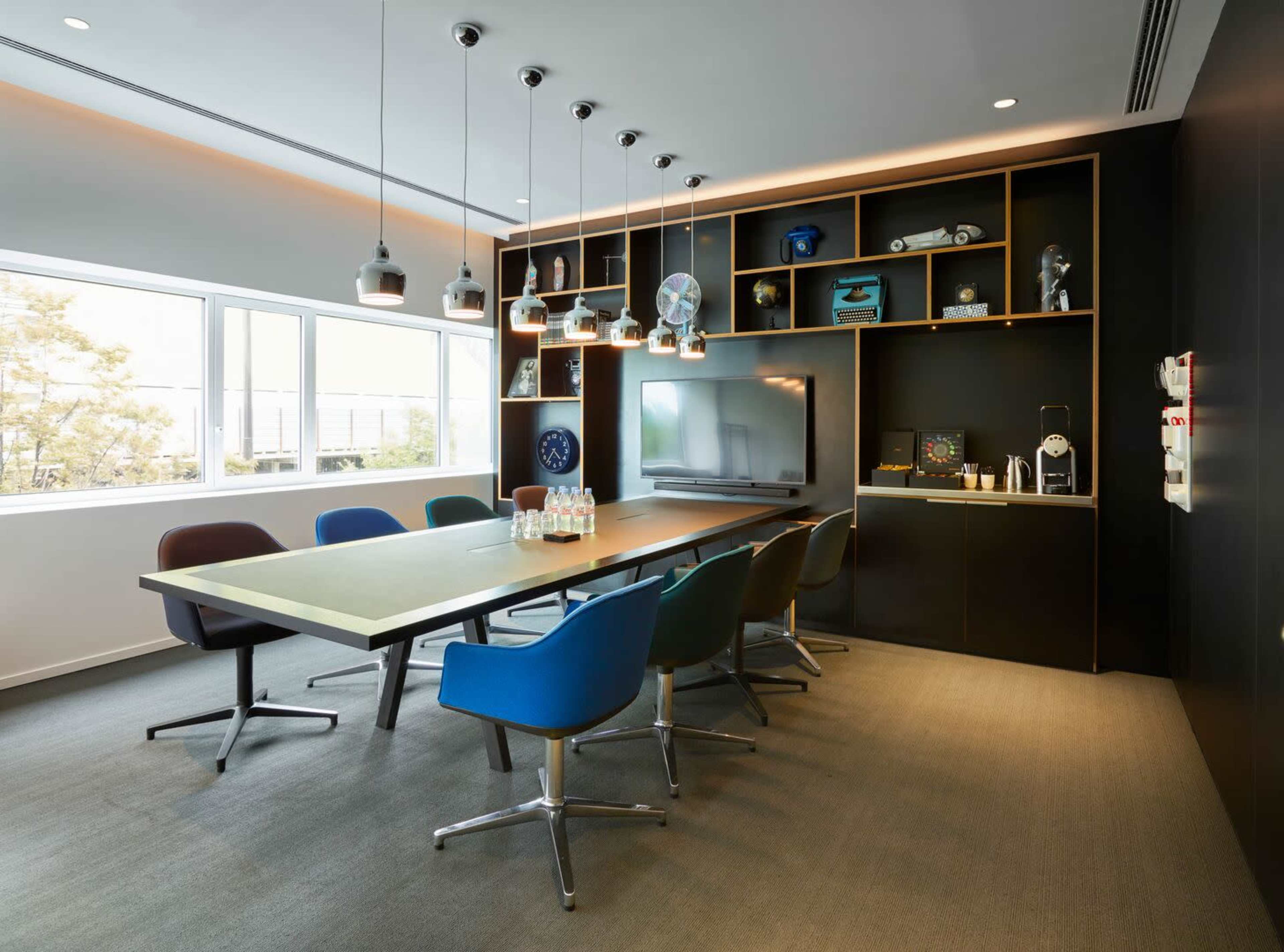 A modern conference room with a long table, assorted colorful chairs, a wall-mounted screen, and shelves displaying various items.