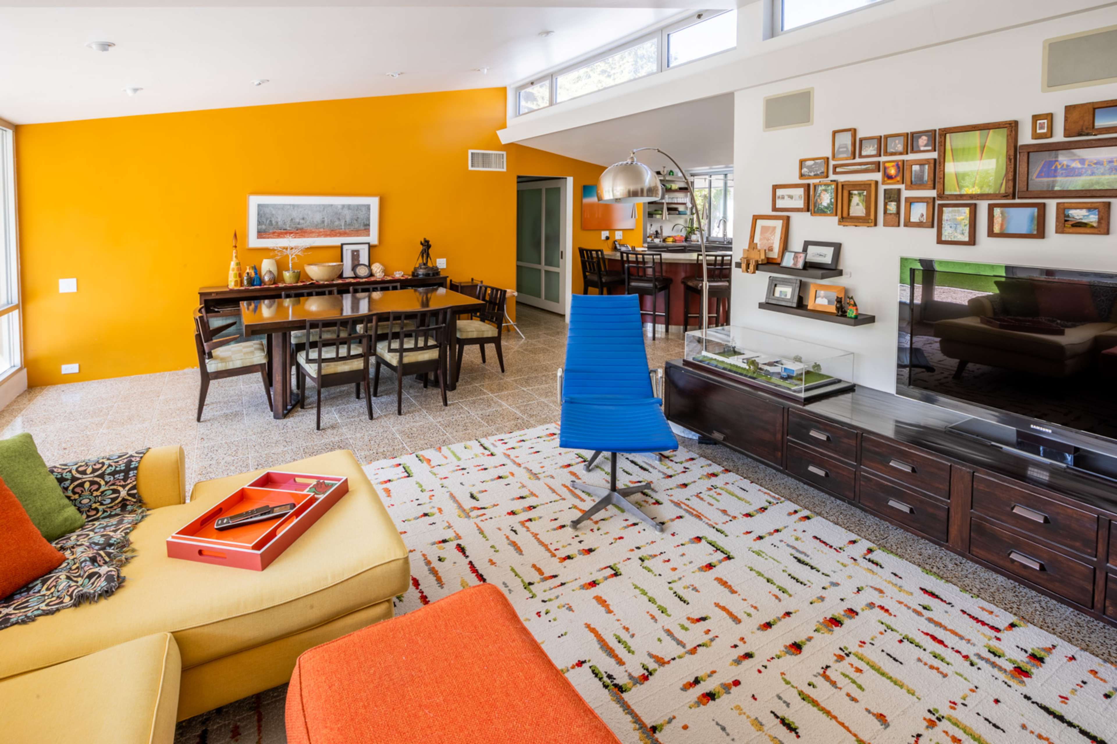 A modern living room with a yellow accent wall, a dining area, and colorful furniture, featuring a large rug with a patterned design.