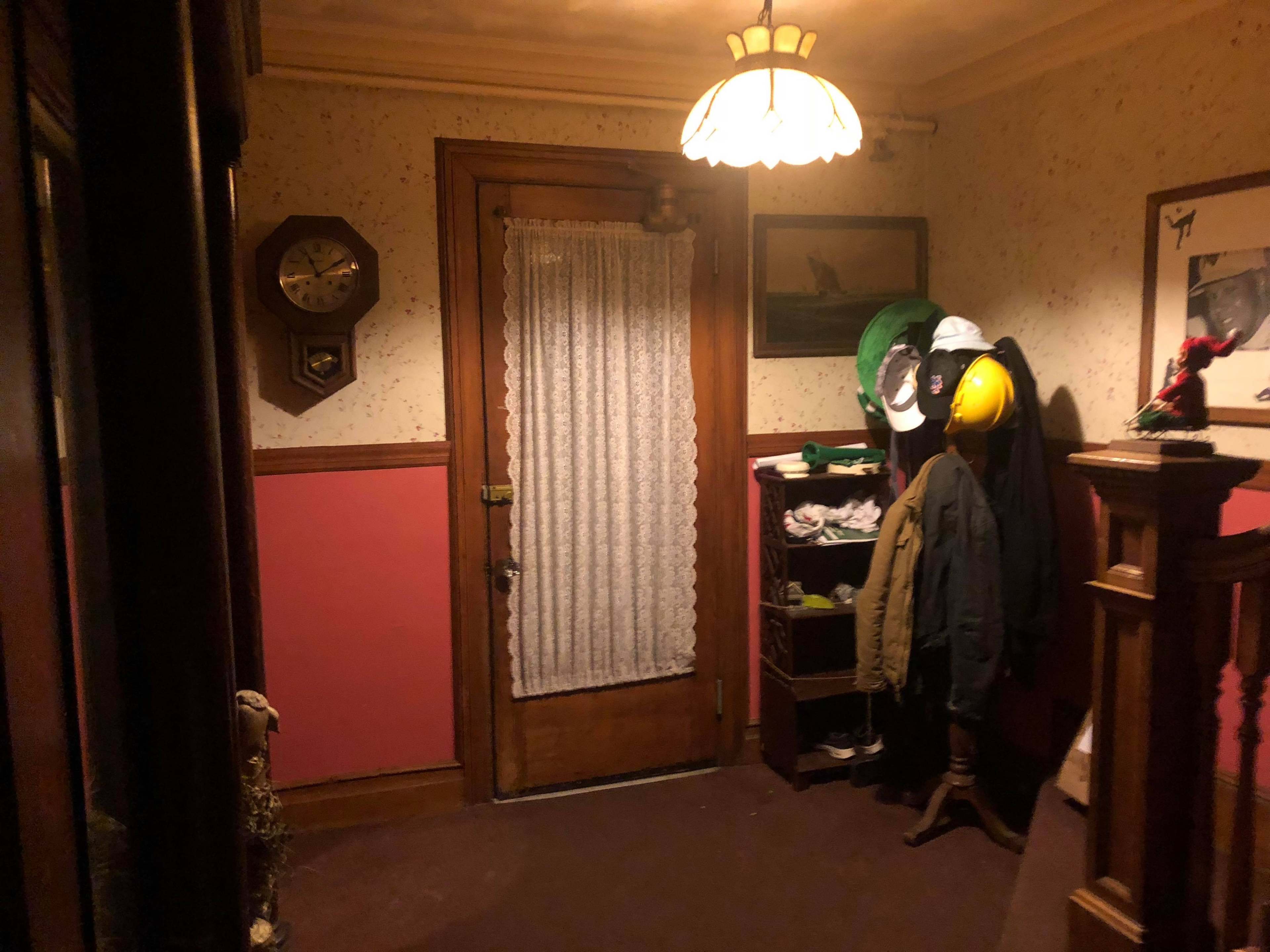 The image shows a dimly lit hallway with a door covered by a lace curtain, adorned with a wall clock, and featuring hooks for hats and coats.