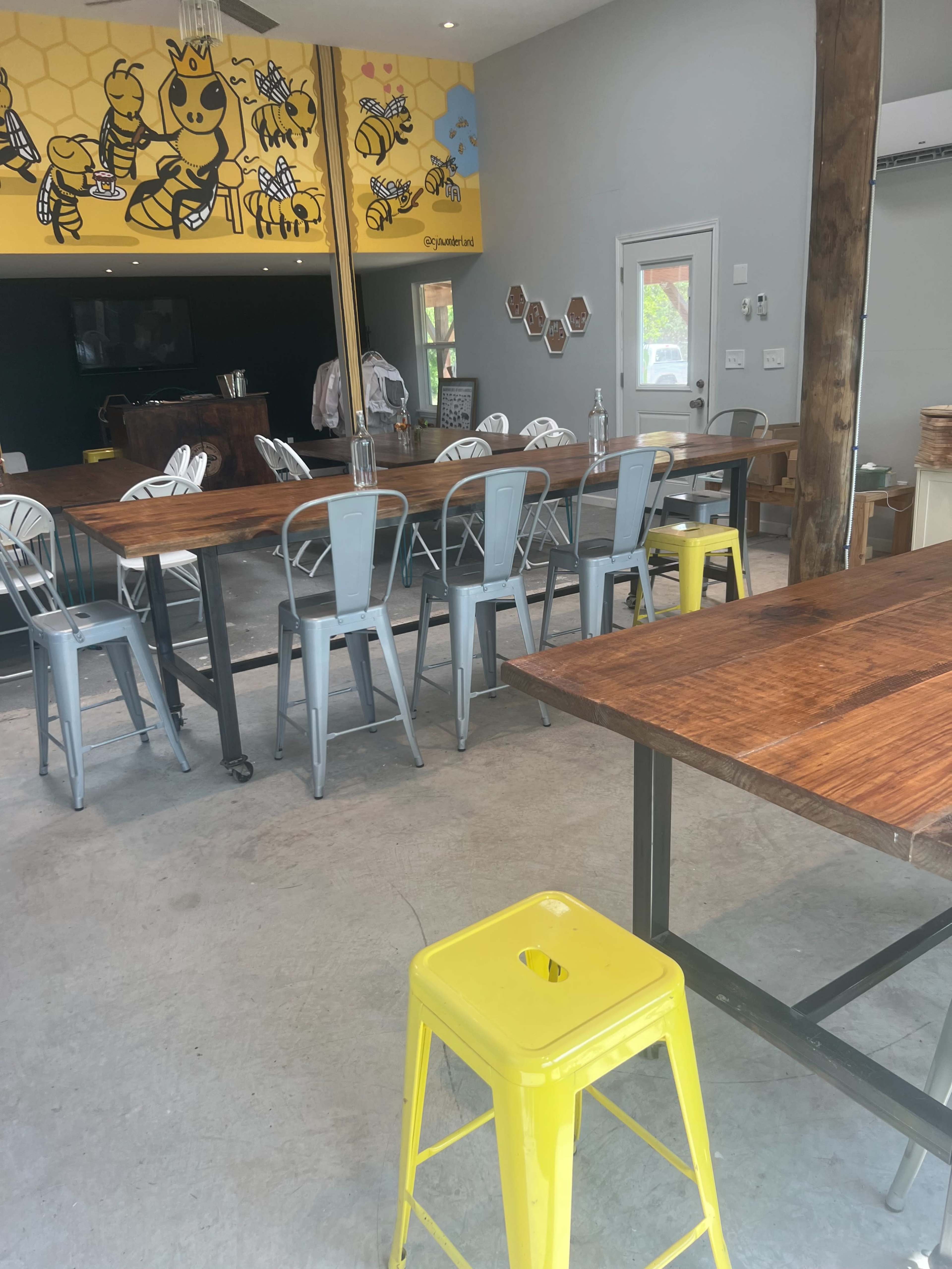 The image shows a modern dining area with metal chairs and a large wooden table, featuring a bee-themed mural on the wall.