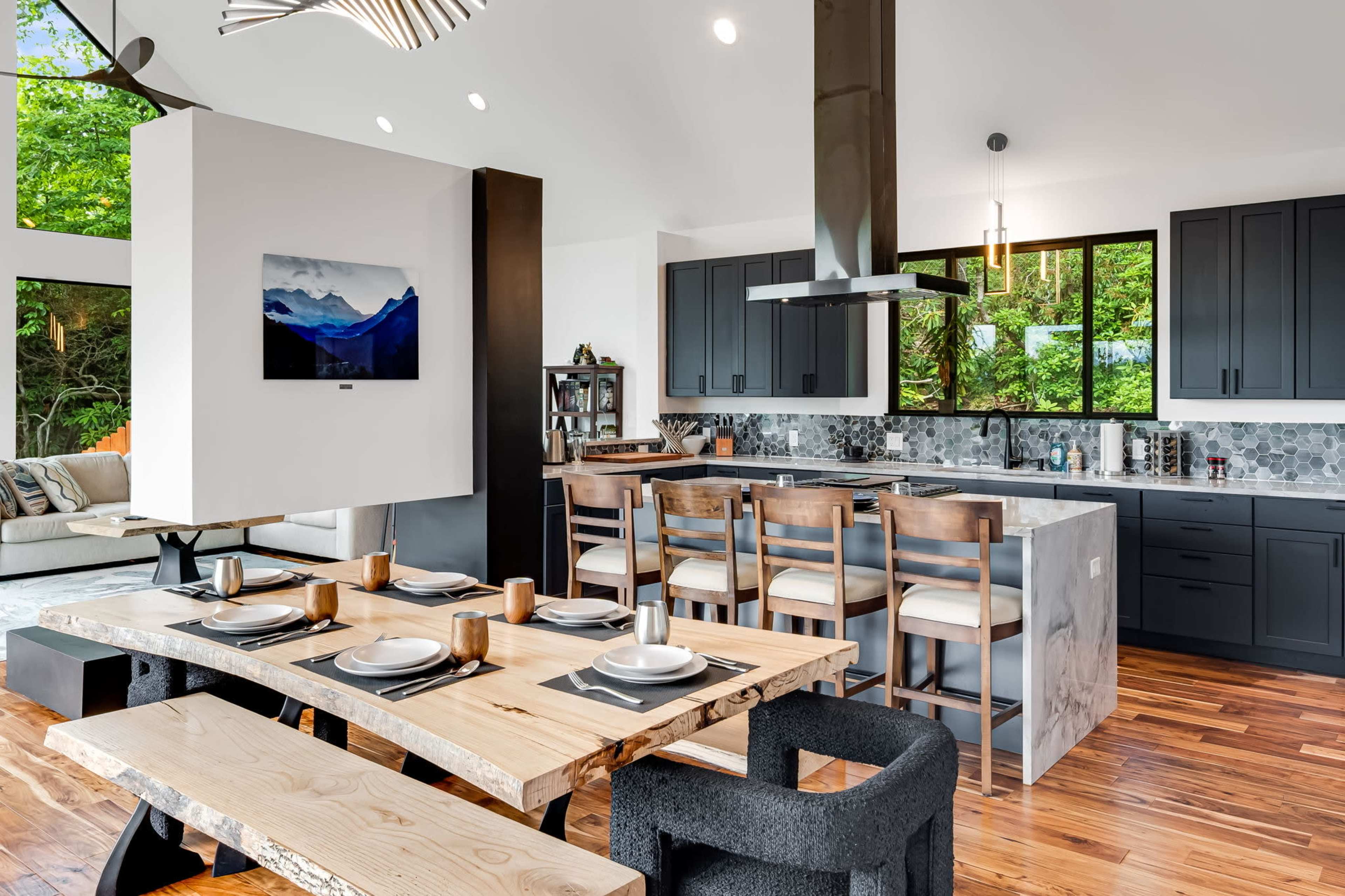 The image shows a modern kitchen with dark cabinetry, a large wooden dining table, and a living area with a couch, all illuminated by natural light from large windows.