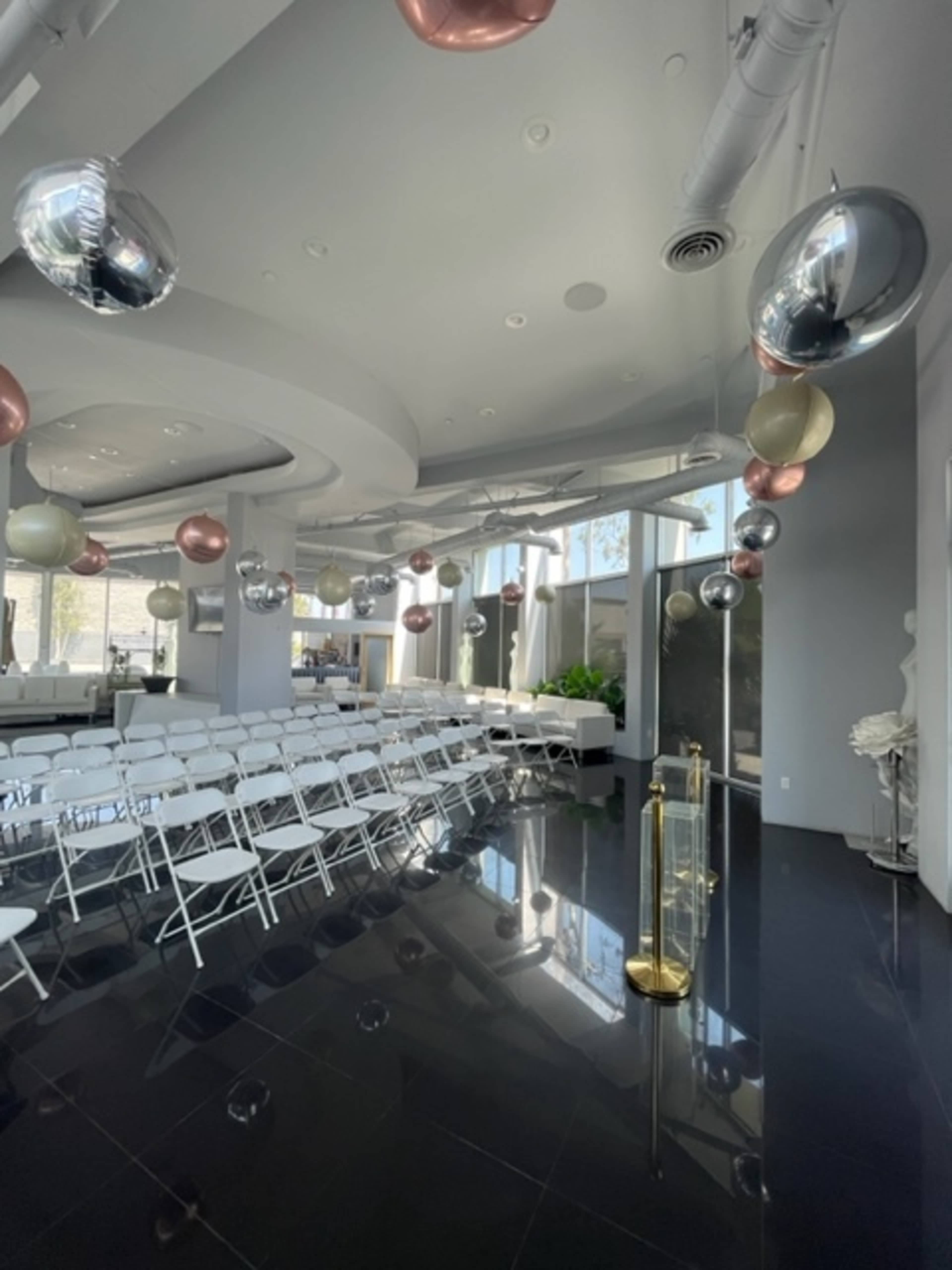 The image shows a modern event space with rows of white chairs, reflective black flooring, and decorative balloons suspended from the ceiling.