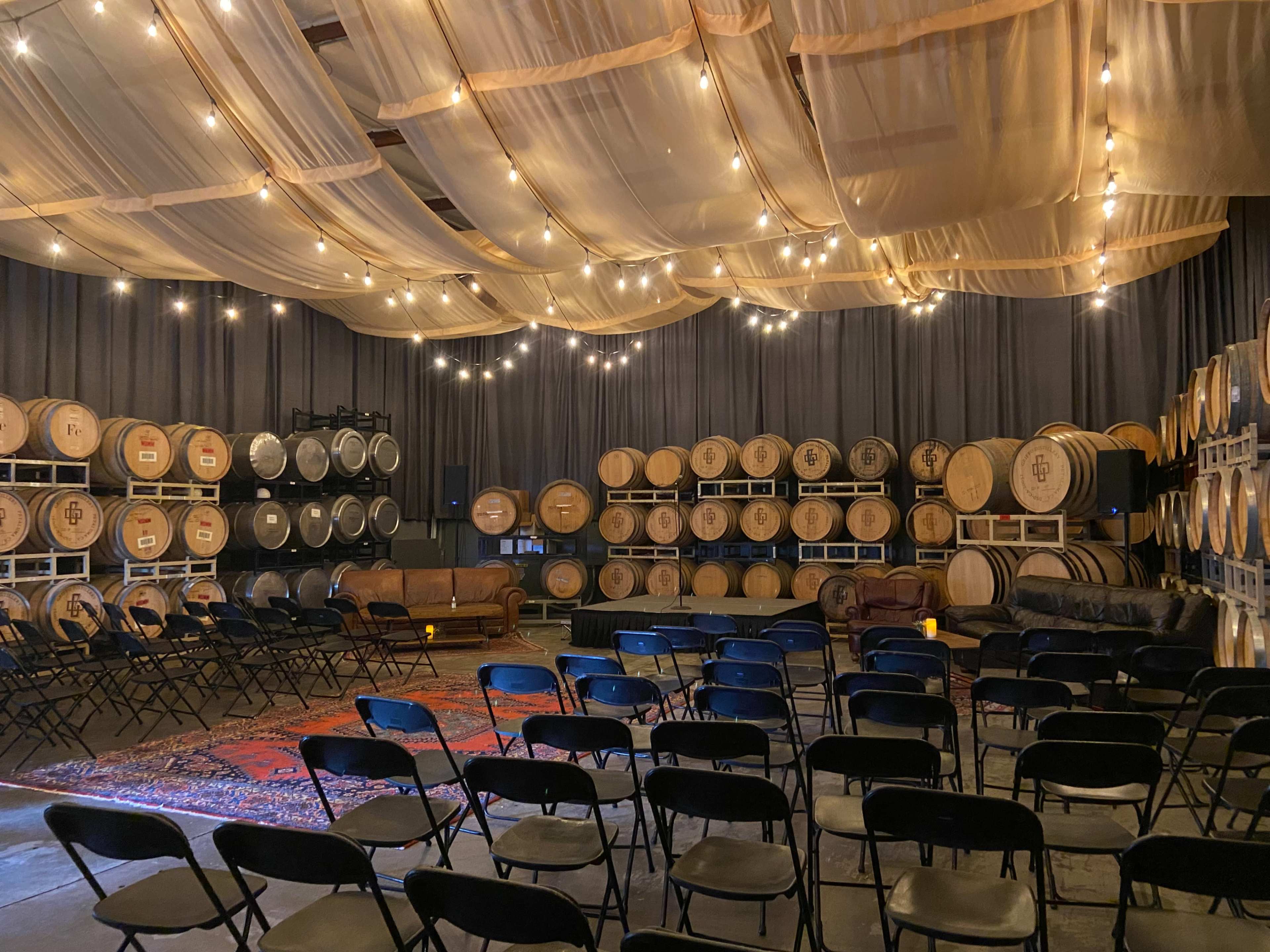 A spacious room with rows of black chairs arranged for an event, surrounded by large wooden barrels and draped fabric above, all illuminated by decorative string lights.