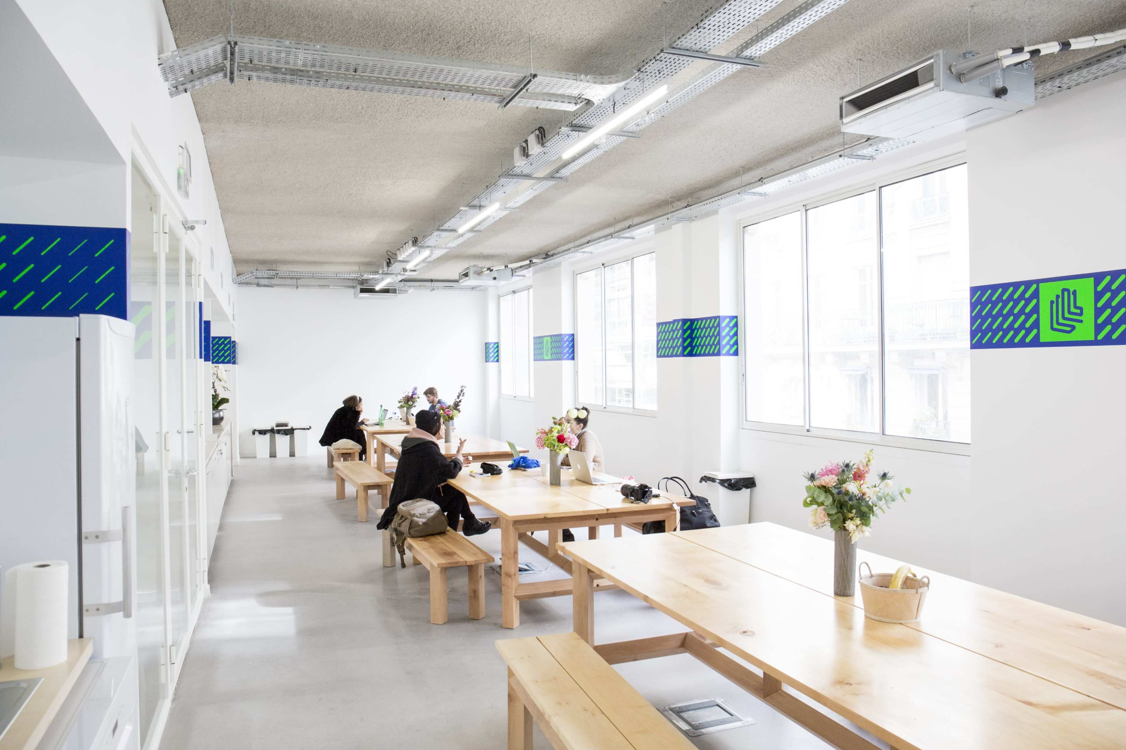 A brightly lit co-working space features long wooden tables with people working quietly and flowers decorating the center.