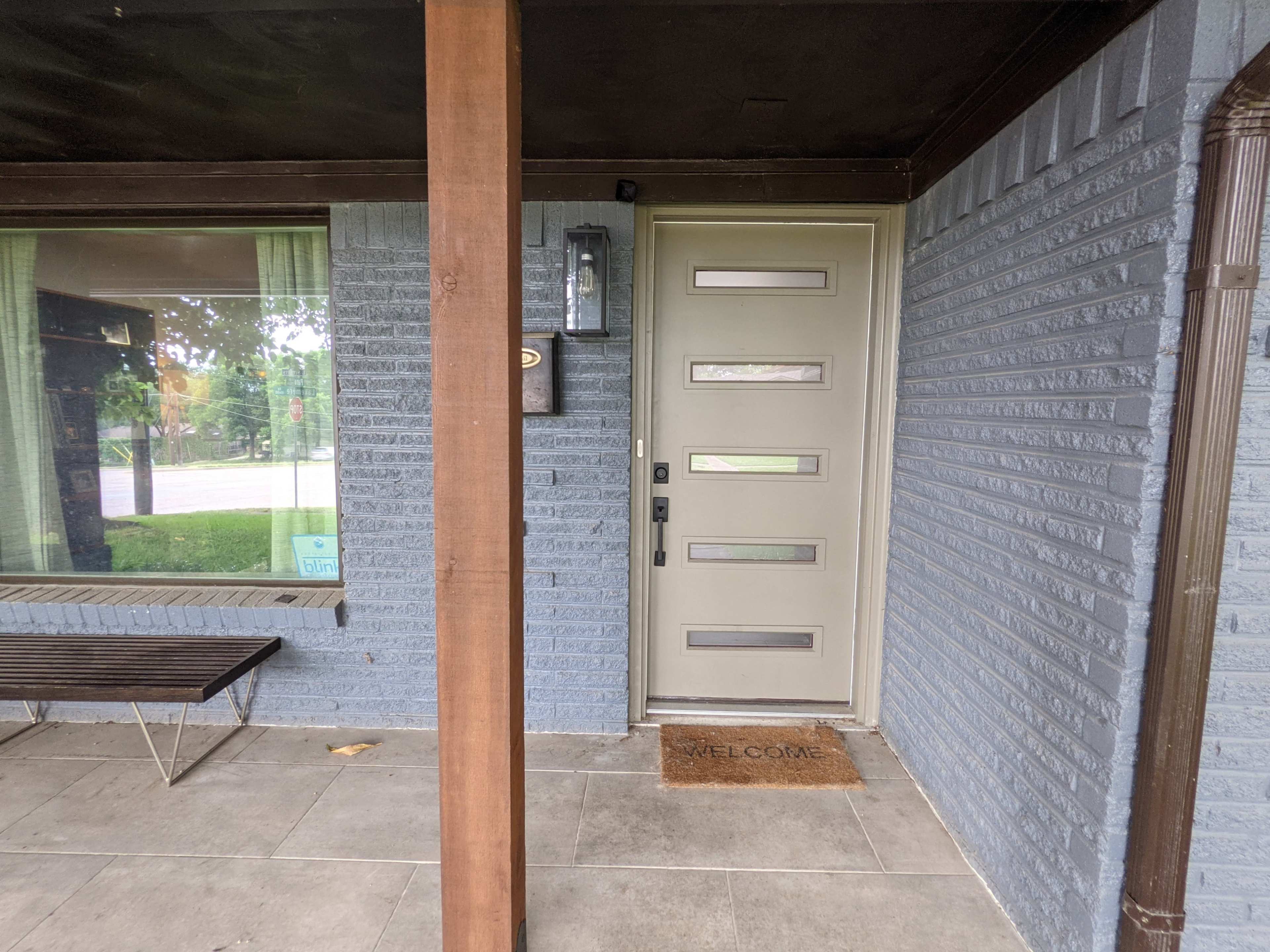 An entryway with a modern door featuring multiple horizontal windows, a welcome mat, and a bench beside a large window.