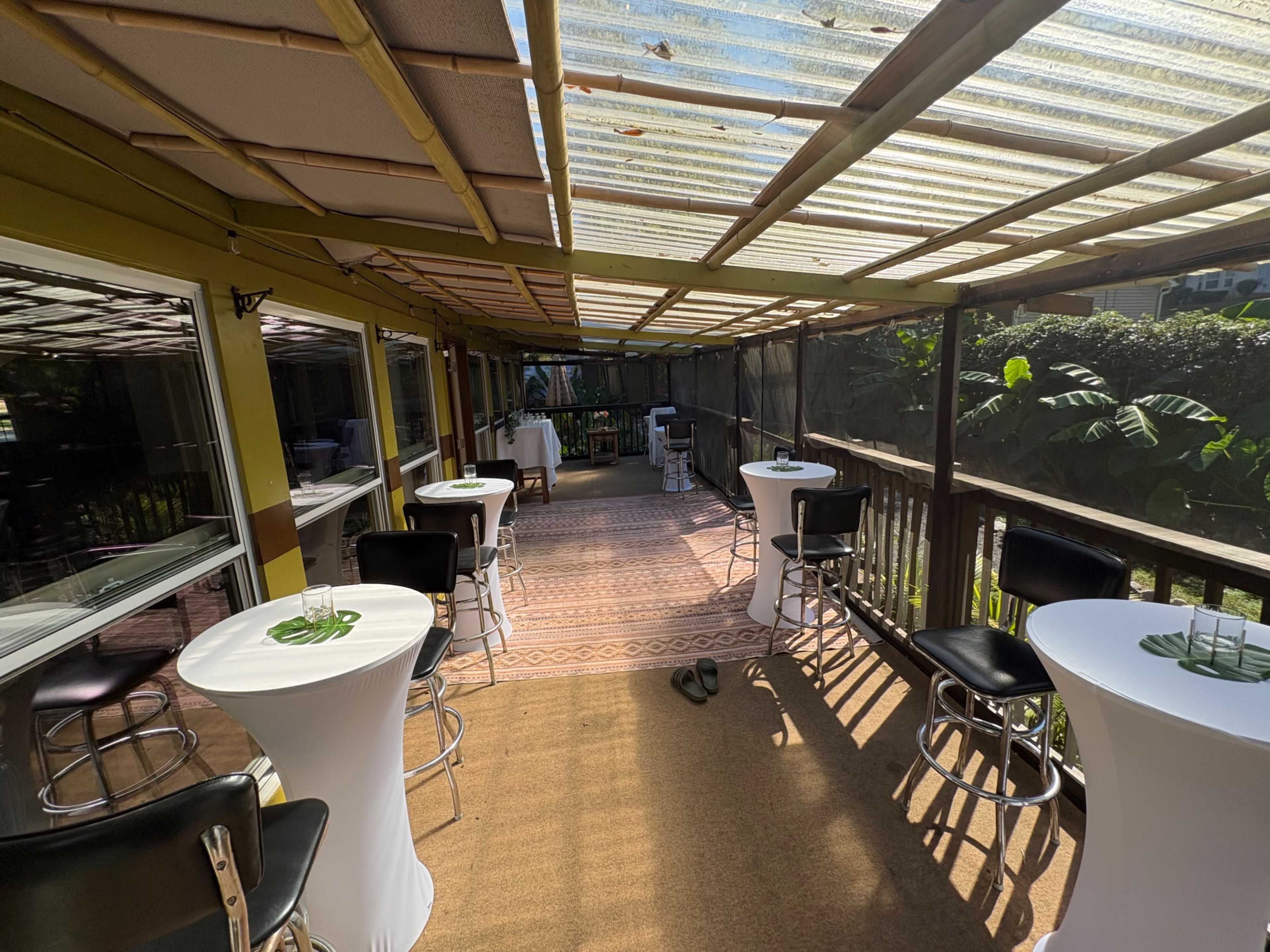 The image shows a well-lit, enclosed outdoor seating area with several tables and chairs, surrounded by greenery.