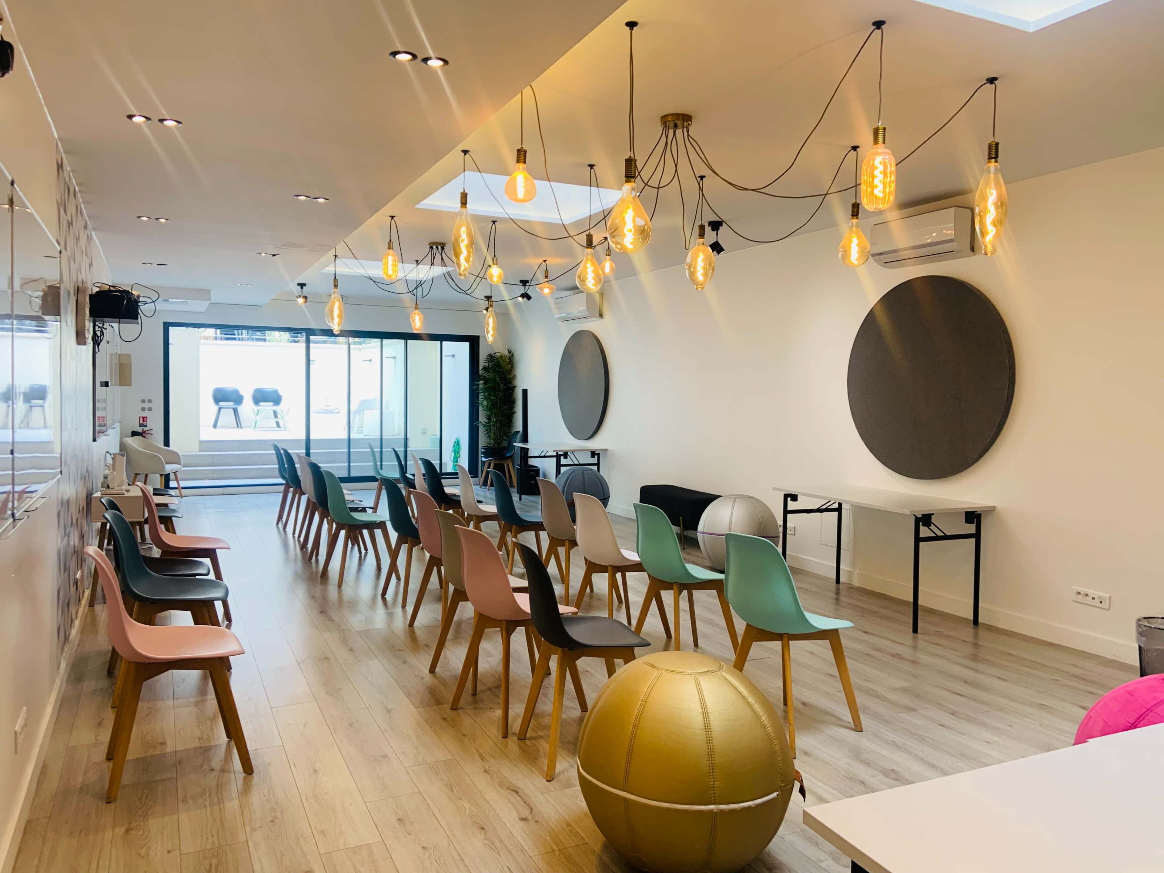 A modern, brightly lit room with rows of colorful chairs facing a glass wall, and decorative pendant lights hanging from the ceiling.