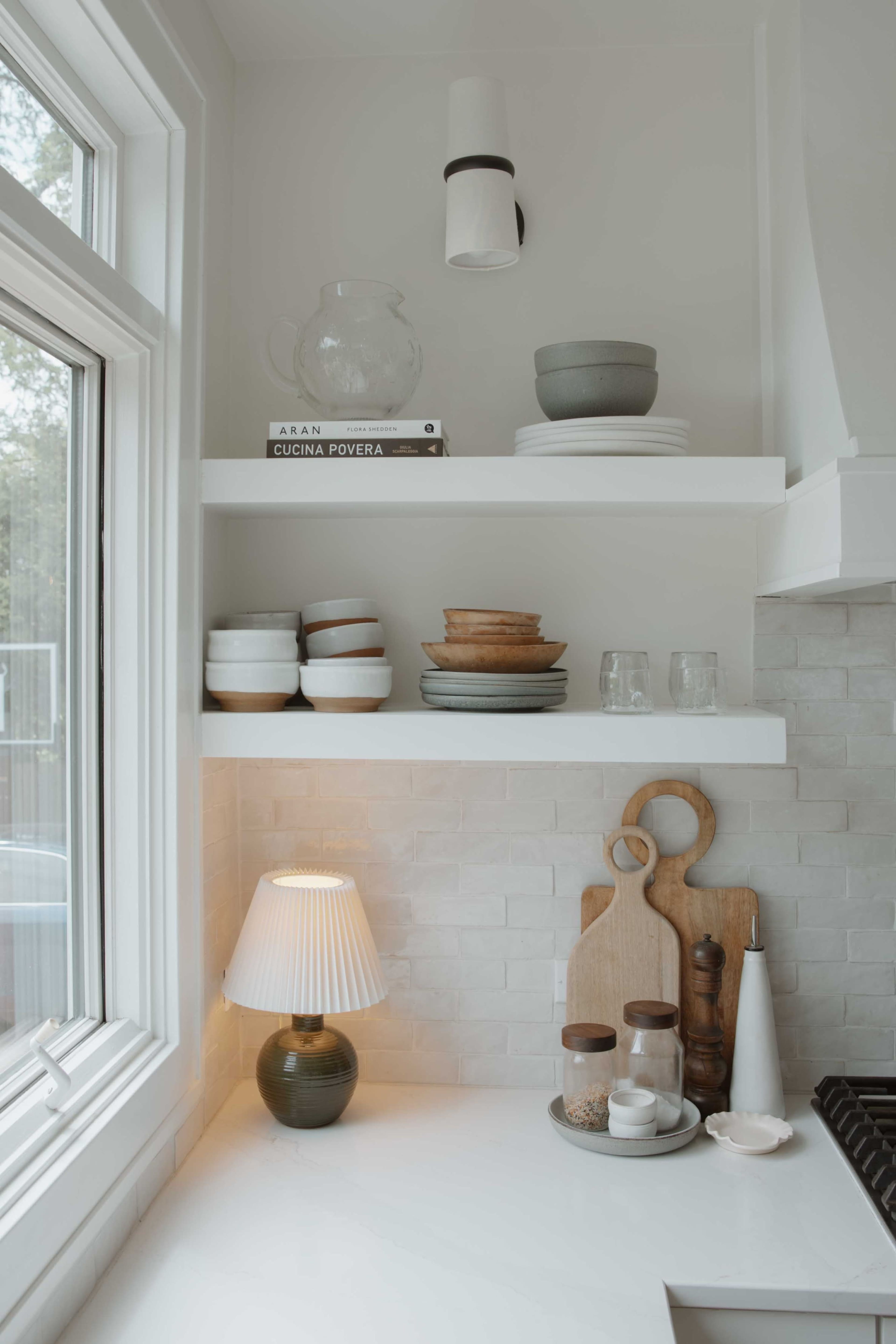 The image shows a well-organized kitchen shelf with various ceramic plates, bowls, and glassware, along with a lamp and wooden cutting boards on a white countertop.