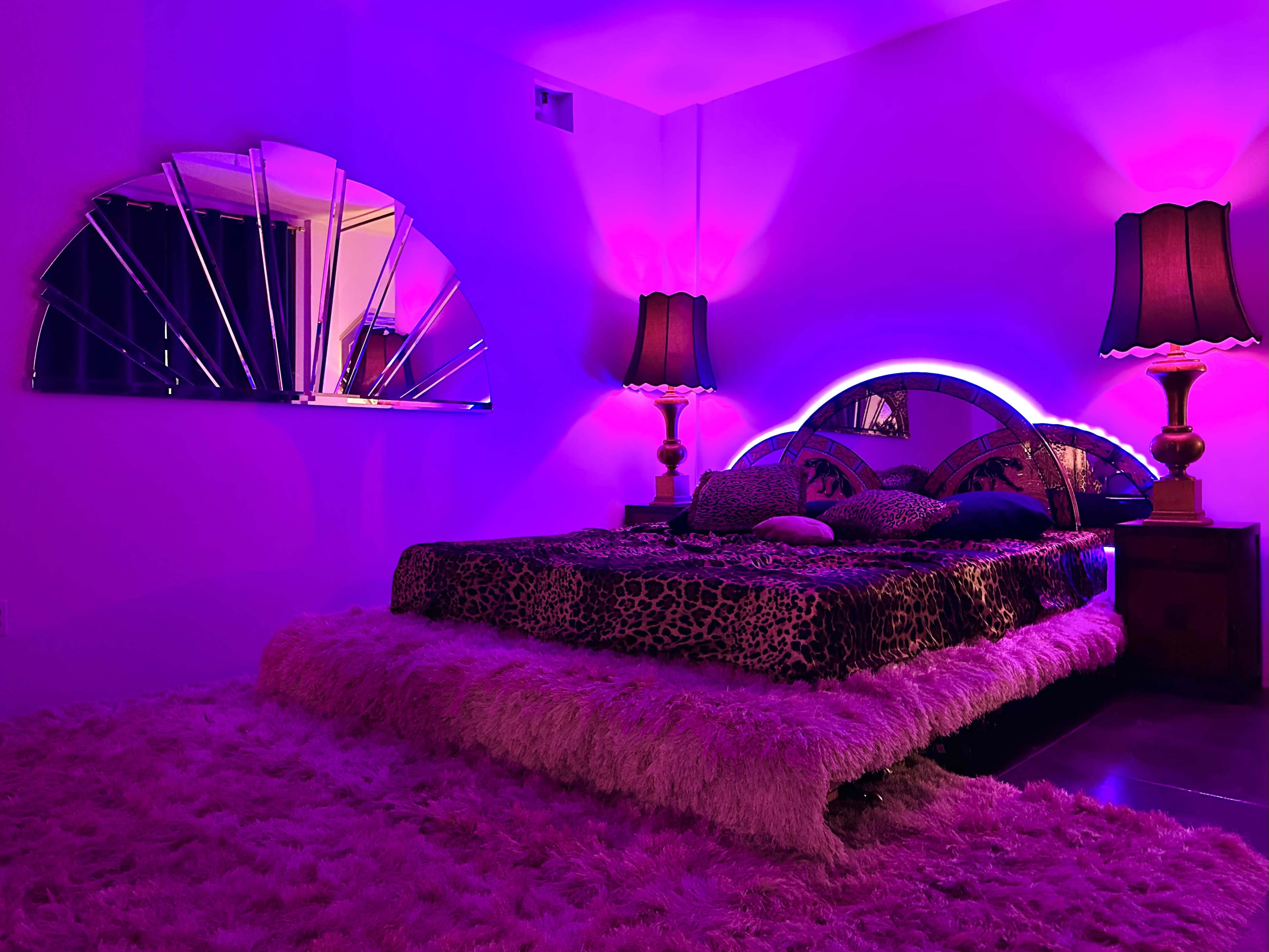A bedroom with a large bed covered in a leopard print comforter, flanked by two lamps and featuring a mirrored wall decoration, all illuminated by purple lighting.
