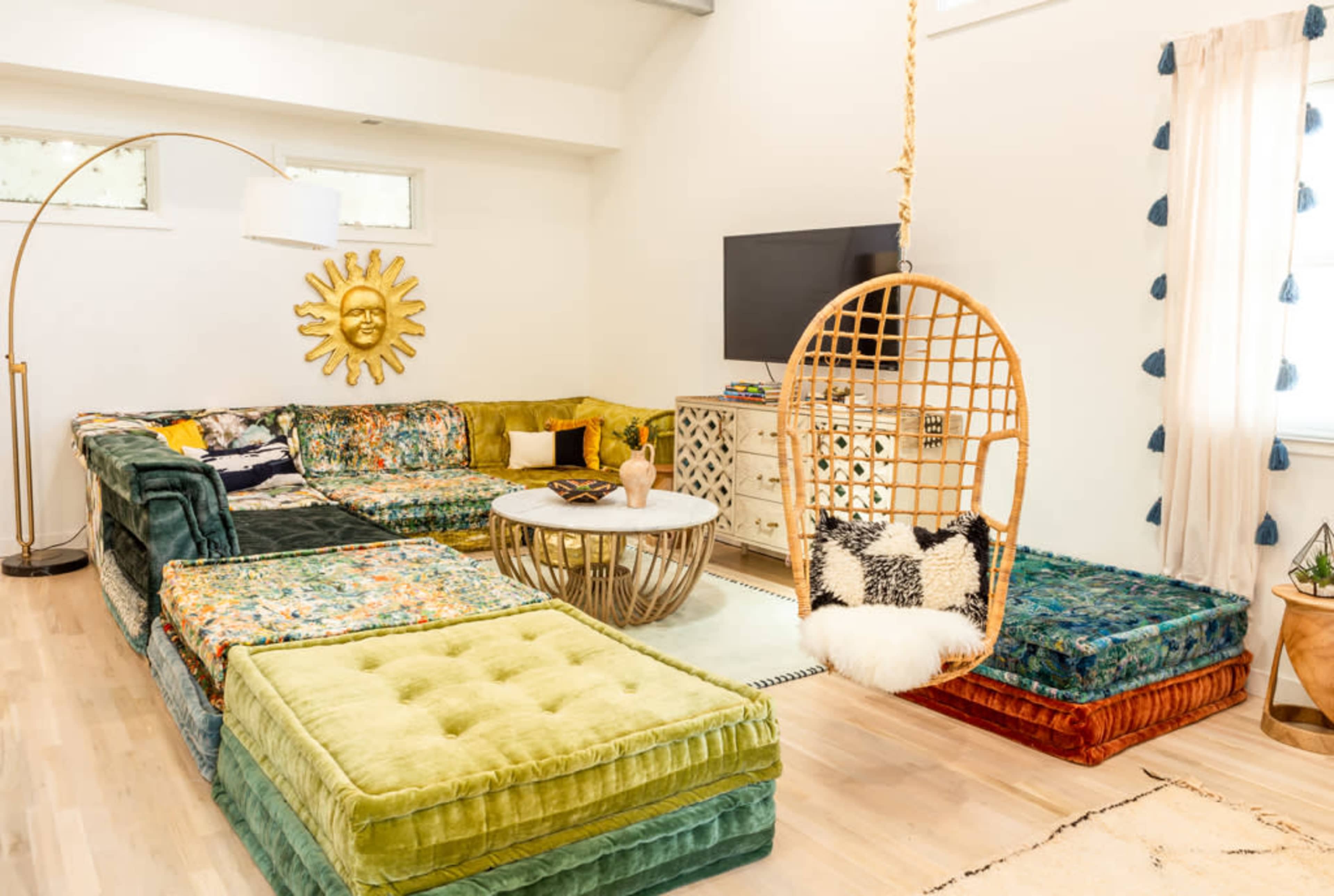 A bright living room featuring a large sectional sofa with colorful cushions, a hanging rattan chair, a round coffee table, and a television against a light-colored wall.