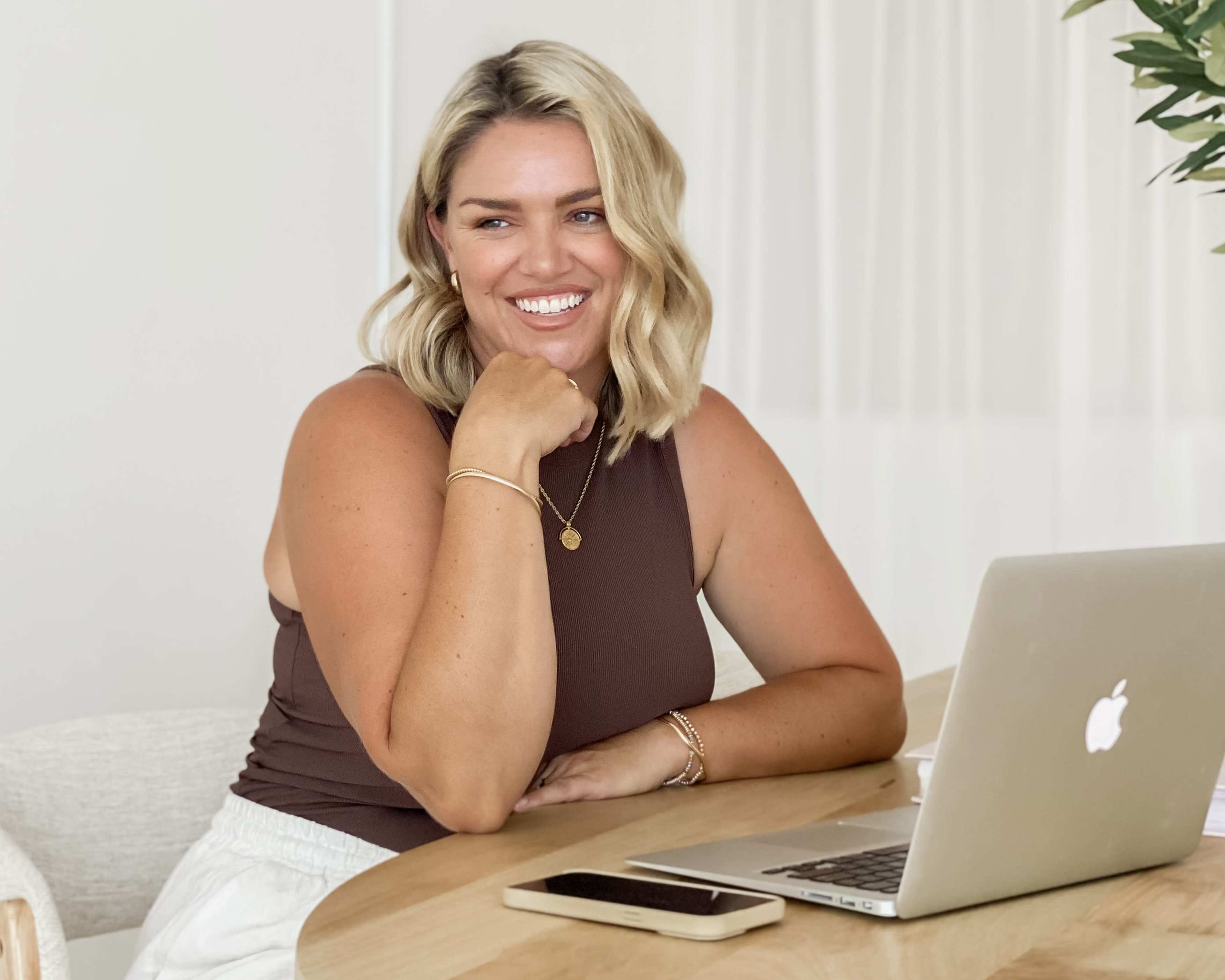 A woman with shoulder-length blonde hair smiles while seated at a wooden table with a laptop and smartphone.
