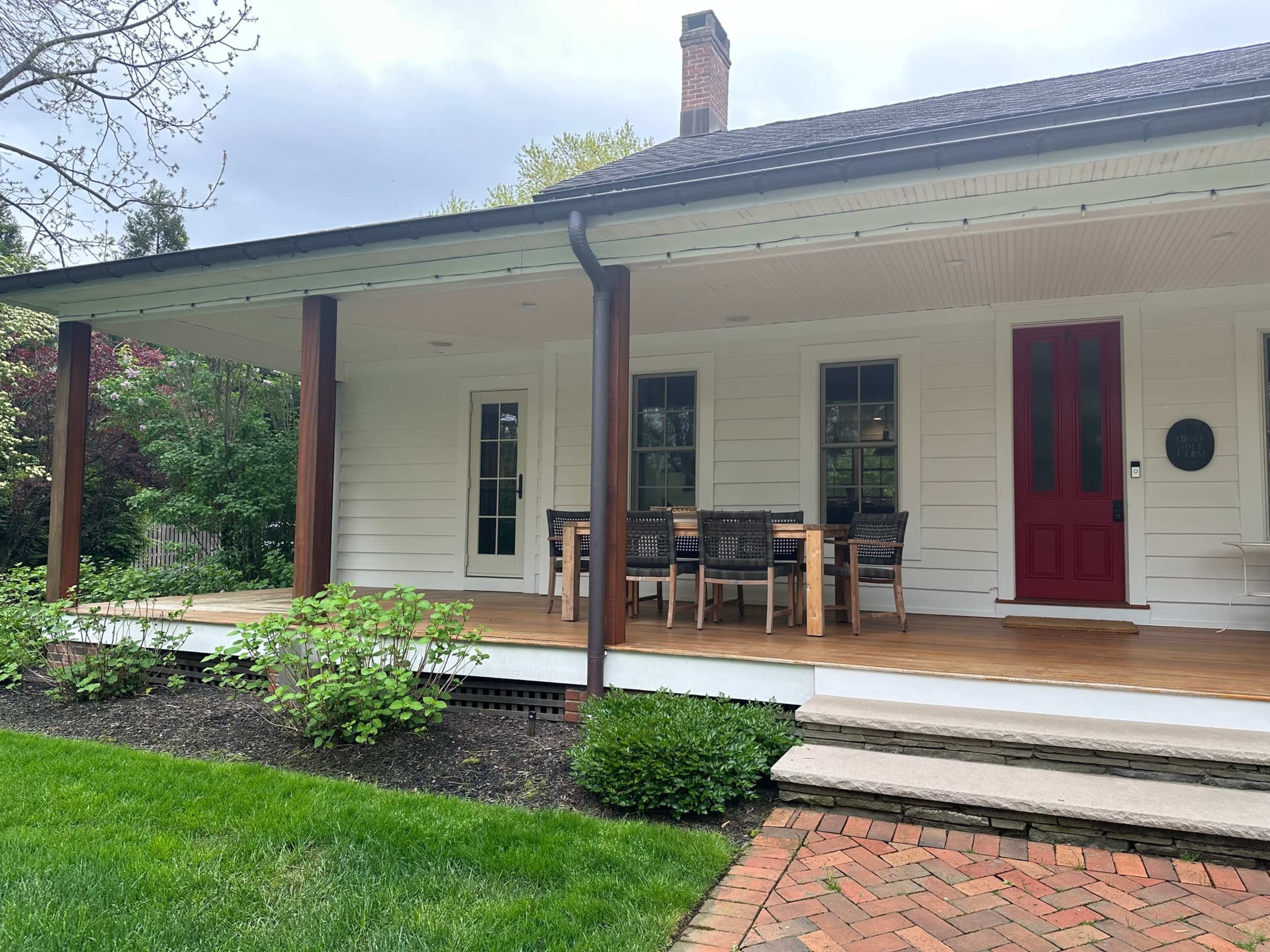The image shows a white house with a red door, featuring a covered porch and a wooden dining table with chairs.