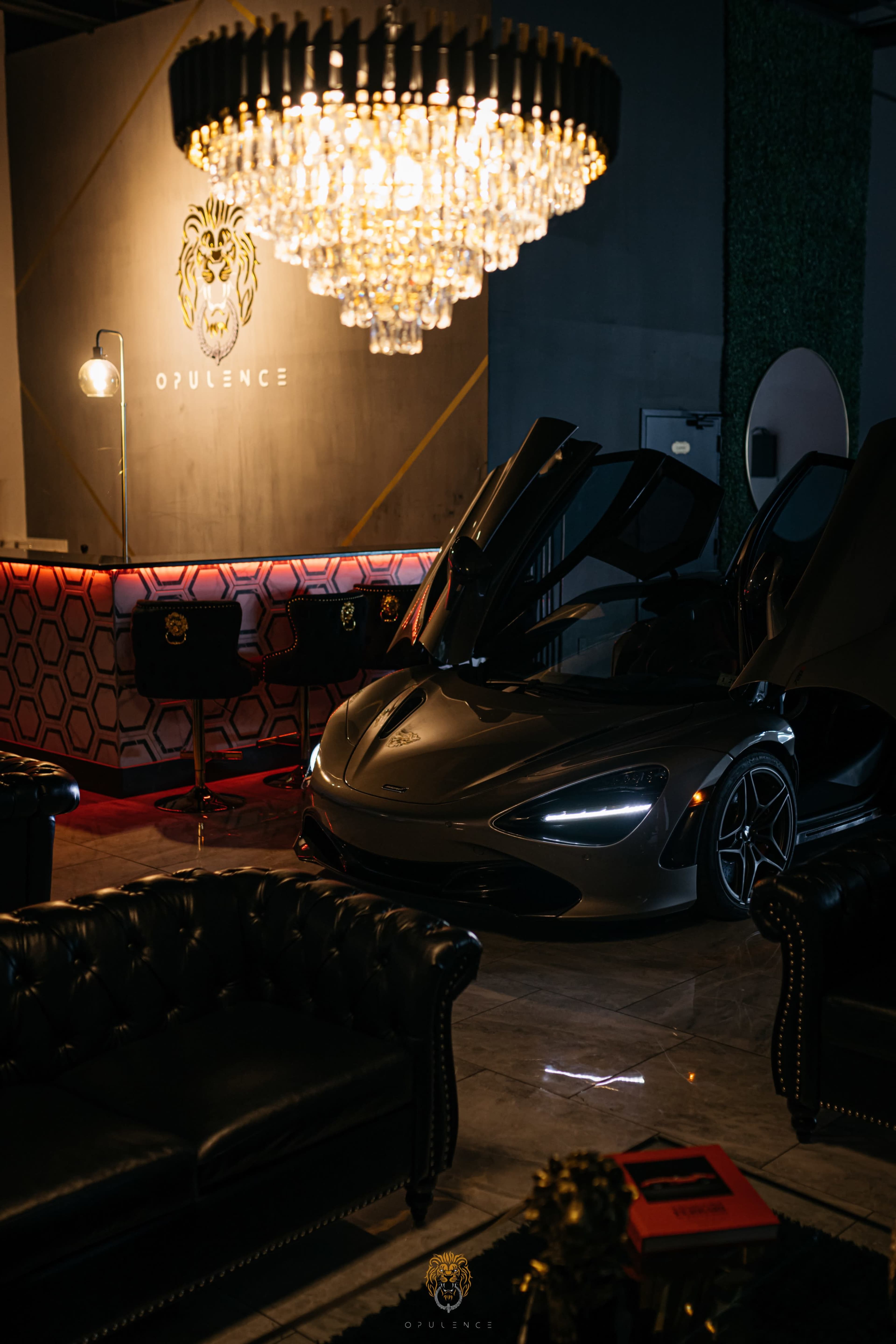 The image features a luxurious lounge area with a parked McLaren 720S sports car, a large chandelier overhead, and elegant seating arrangements.