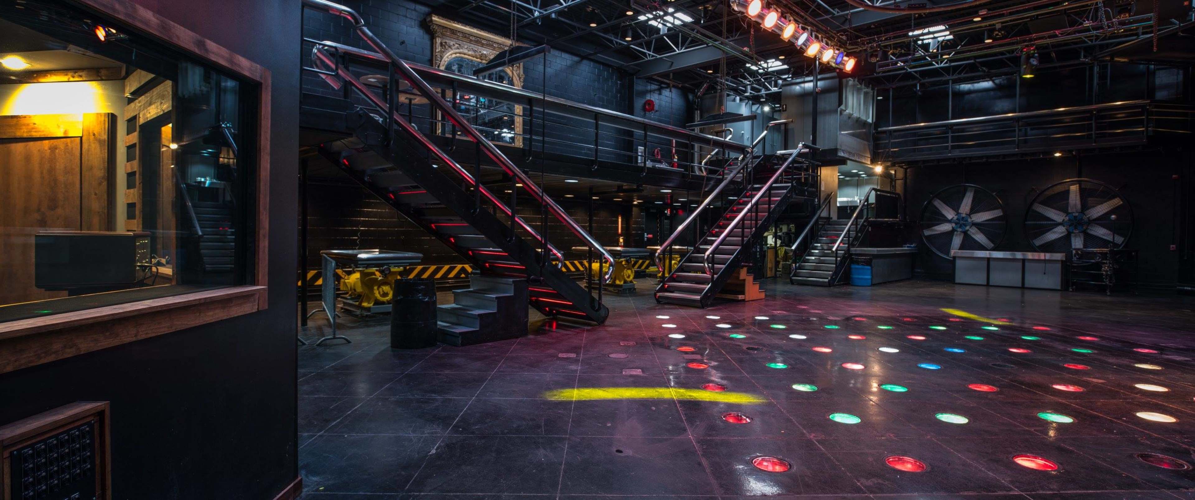 A spacious performance venue with a dark interior, featuring two sets of metal stairs, disco lights on the floor, and industrial elements like large fans.