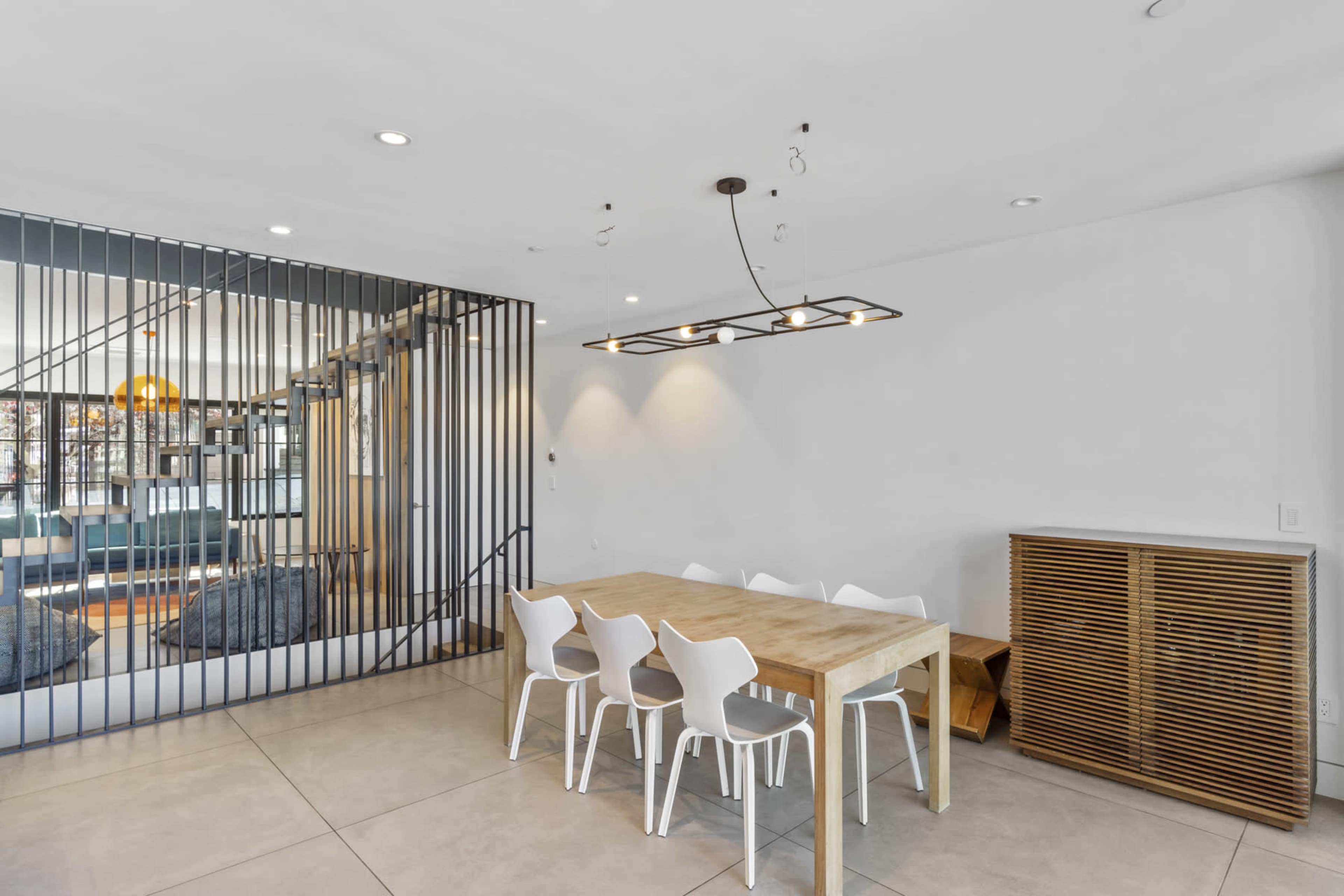 The image shows a modern dining area featuring a wooden table surrounded by white chairs, with a partition of black vertical bars in the background.