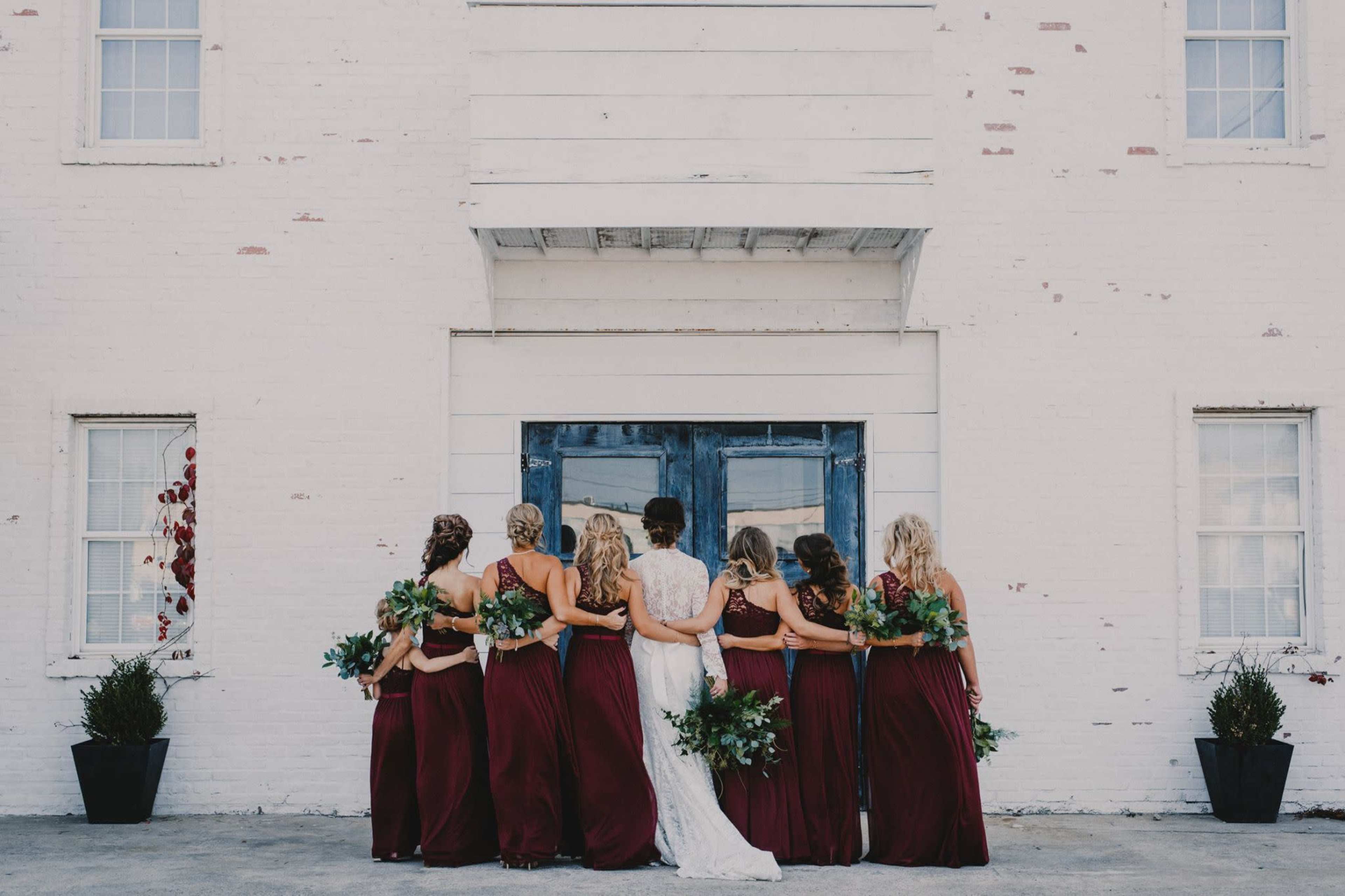 A bride and her bridesmaids stand together in matching maroon dresses in front of a white building with blue doors.