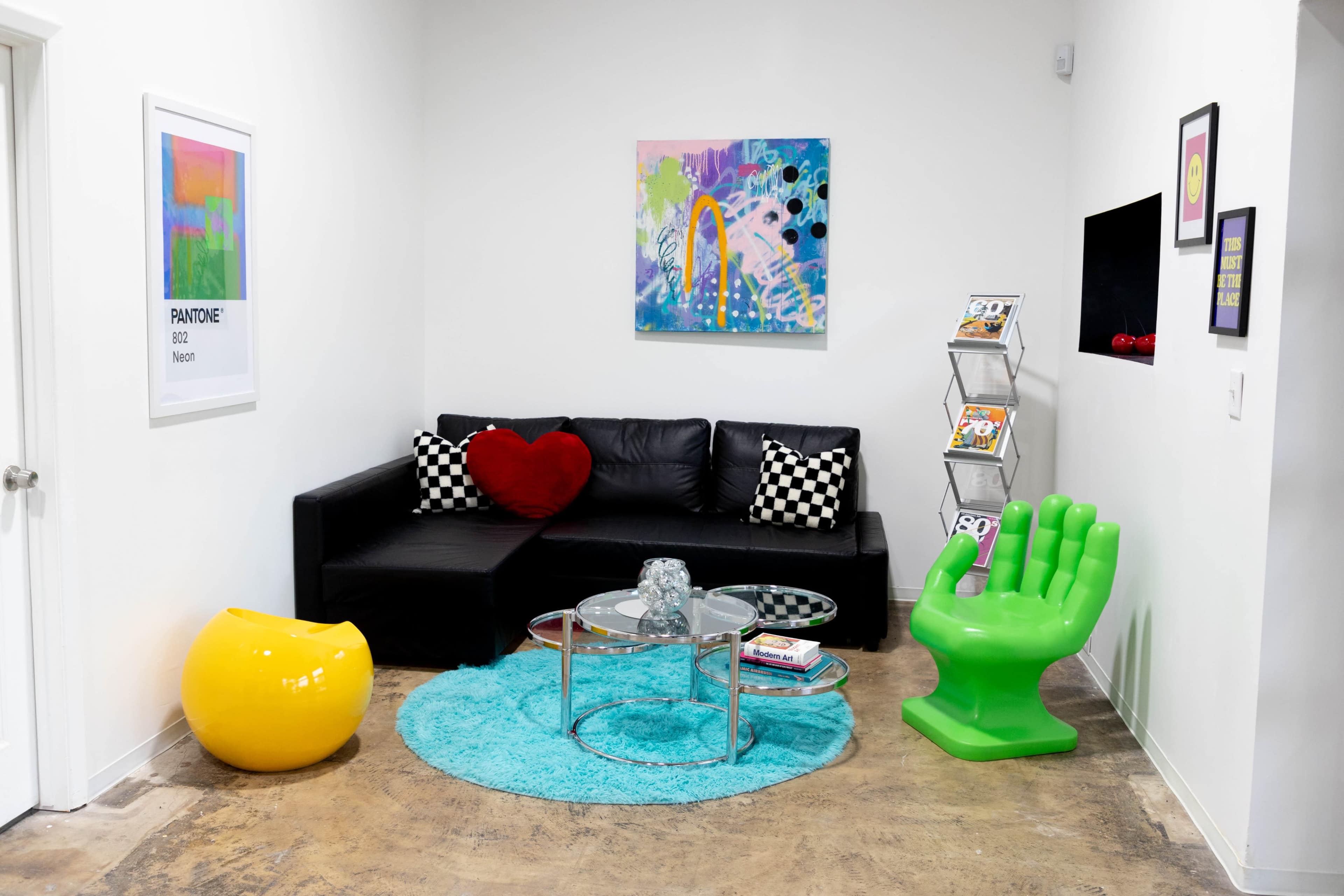 A small, modern living space features a black sectional sofa with a red heart-shaped cushion, a glass coffee table, a bright yellow decor piece, a green hand-shaped chair, and a blue rug on a concrete floor.