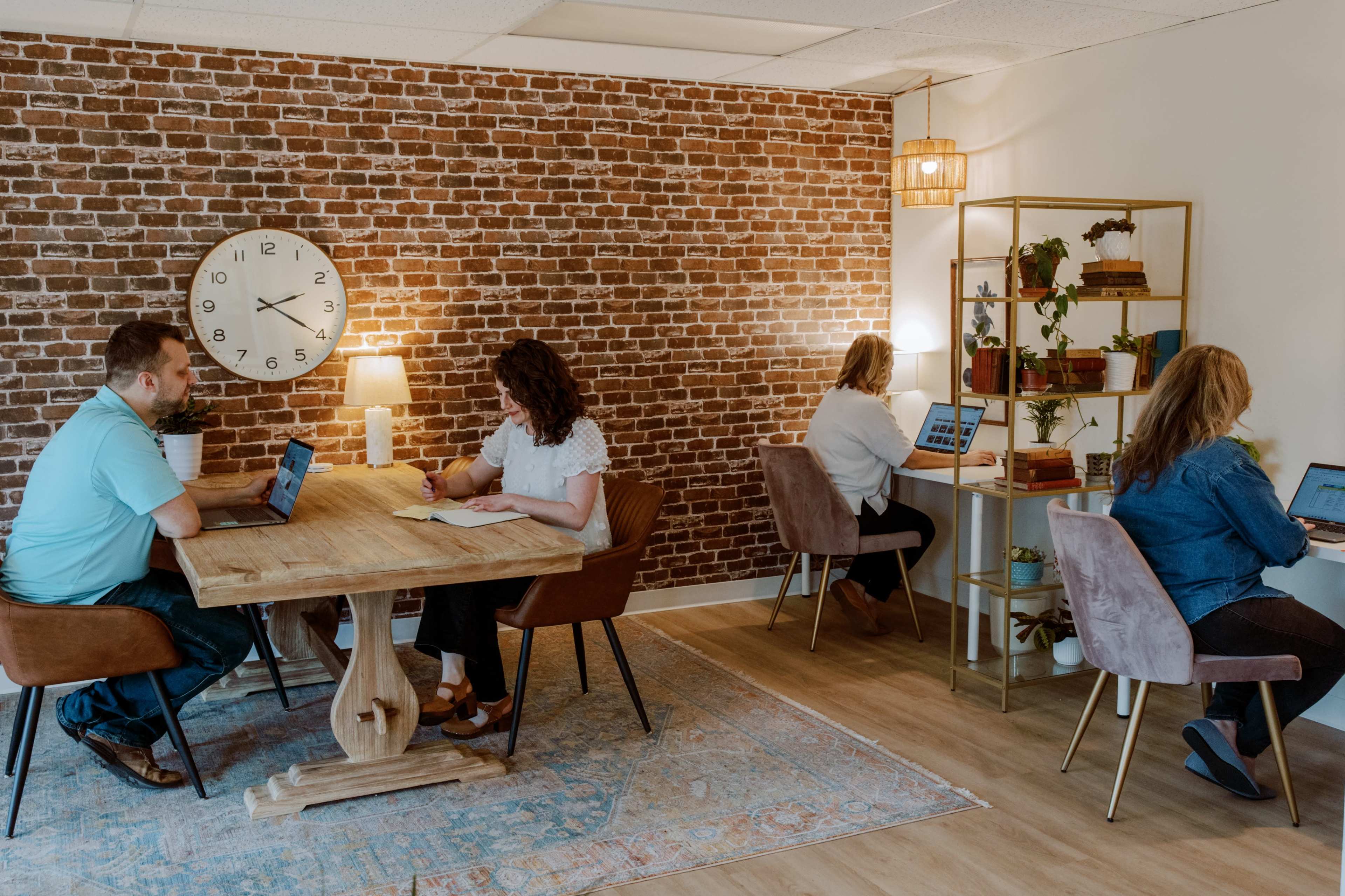 The image shows a modern workspace with a rustic brick wall, featuring four people engaged in work at a wooden table and separate desks, surrounded by plants and decorative lighting.