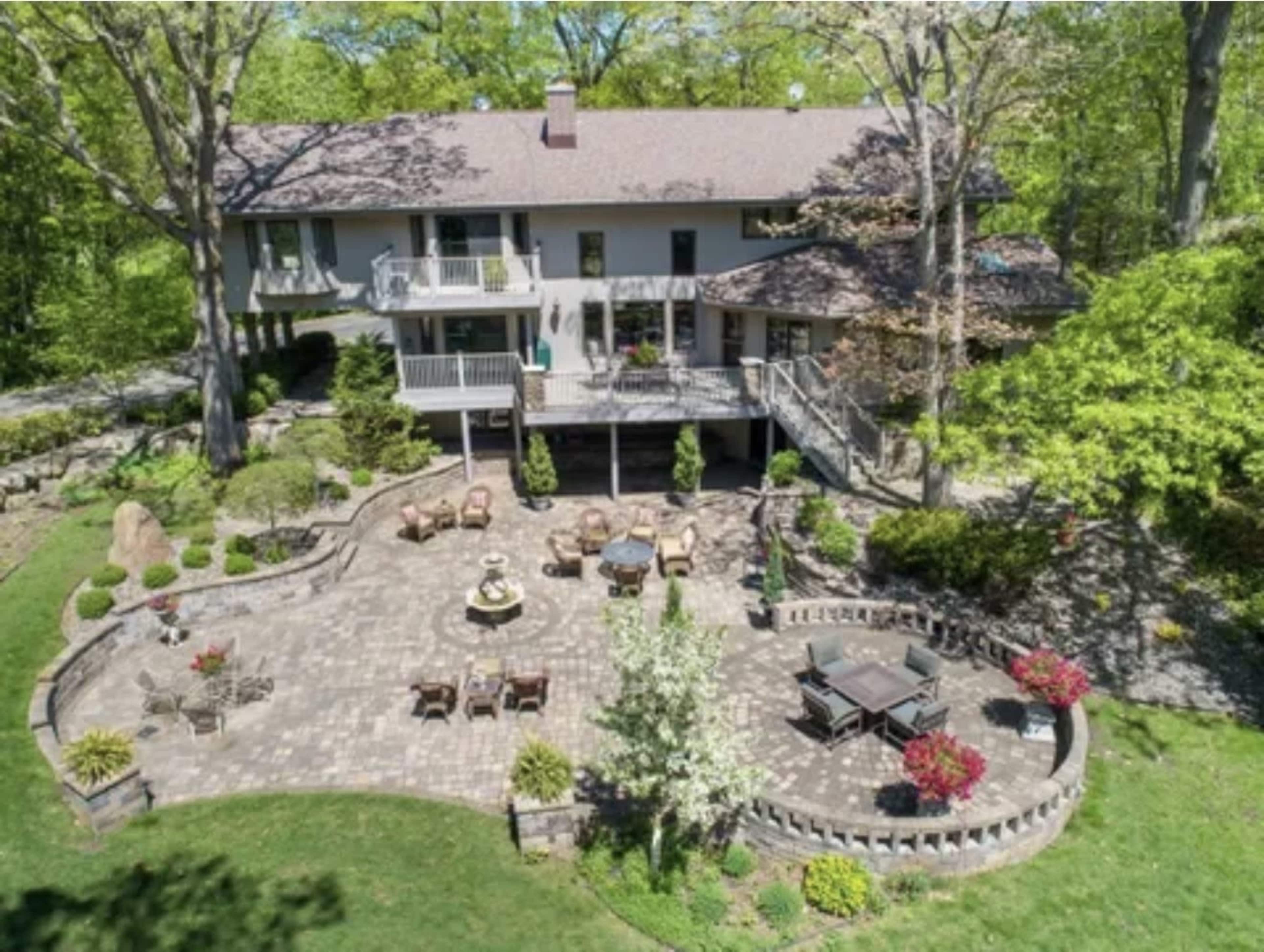 The image shows a spacious home with a multi-level deck overlooking a landscaped backyard featuring a brick patio, seating areas, and a fountain.