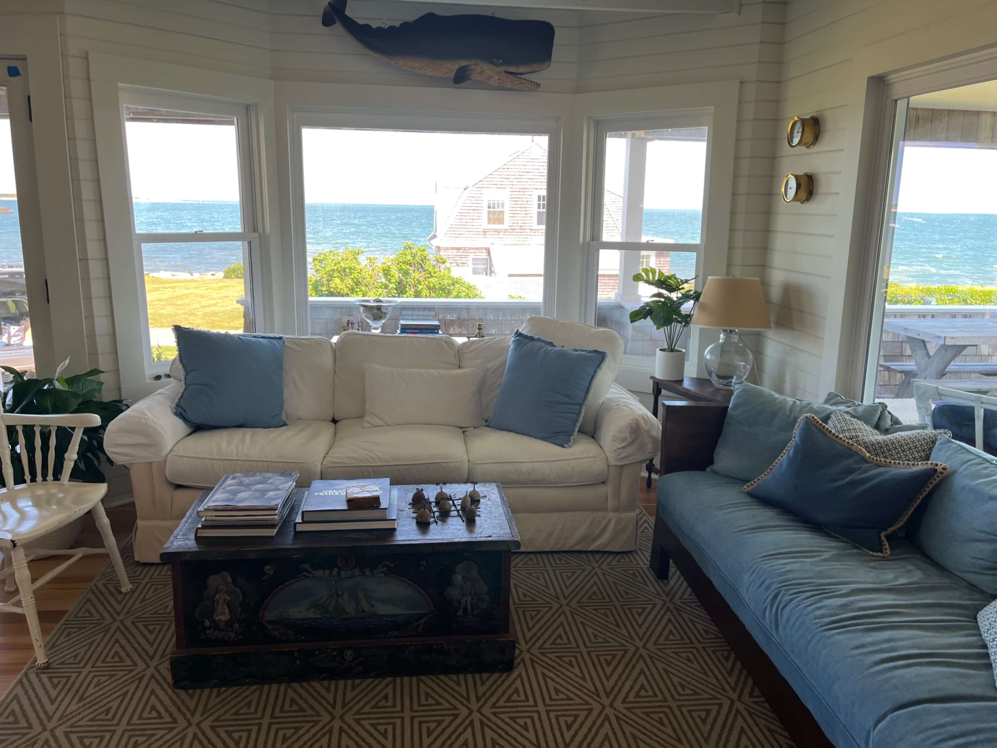 A bright living room features a white couch with blue accents, a dark wooden coffee table, and windows overlooking the ocean.