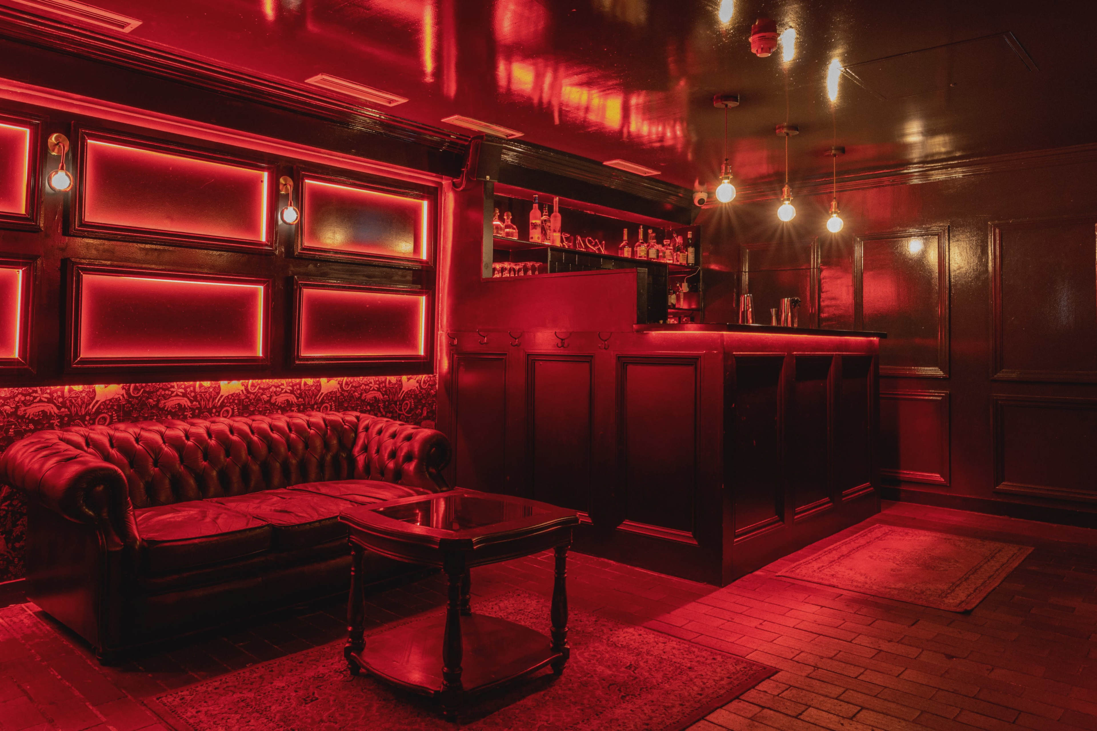 A dimly lit bar interior with red-tinted lighting, featuring a leather couch and a small wooden coffee table in front of a bar counter.