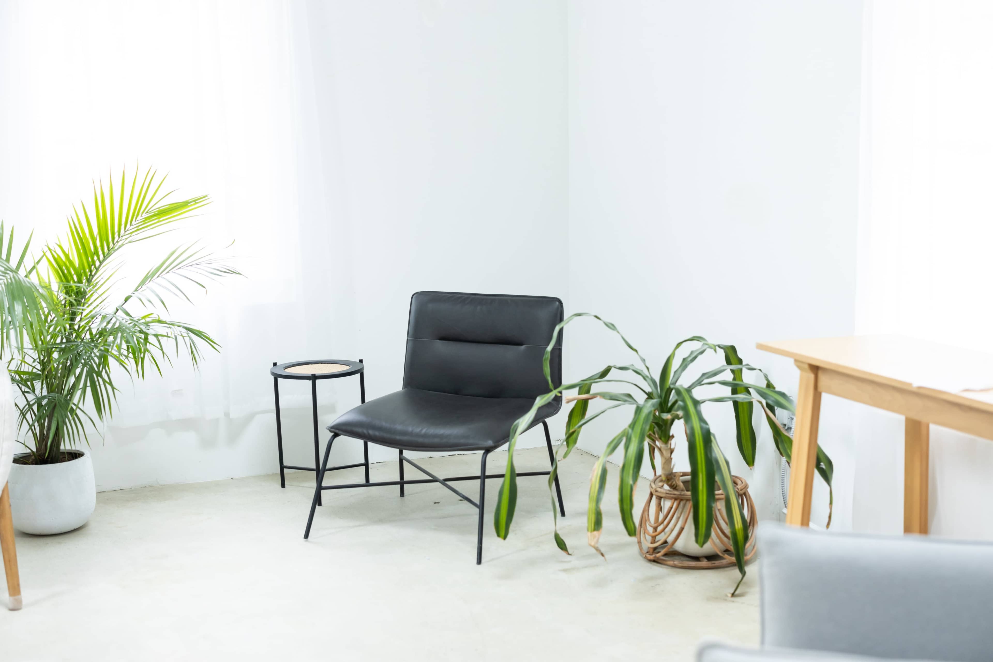 A black lounge chair sits next to a small round side table and a potted plant in a well-lit, minimalistic room.