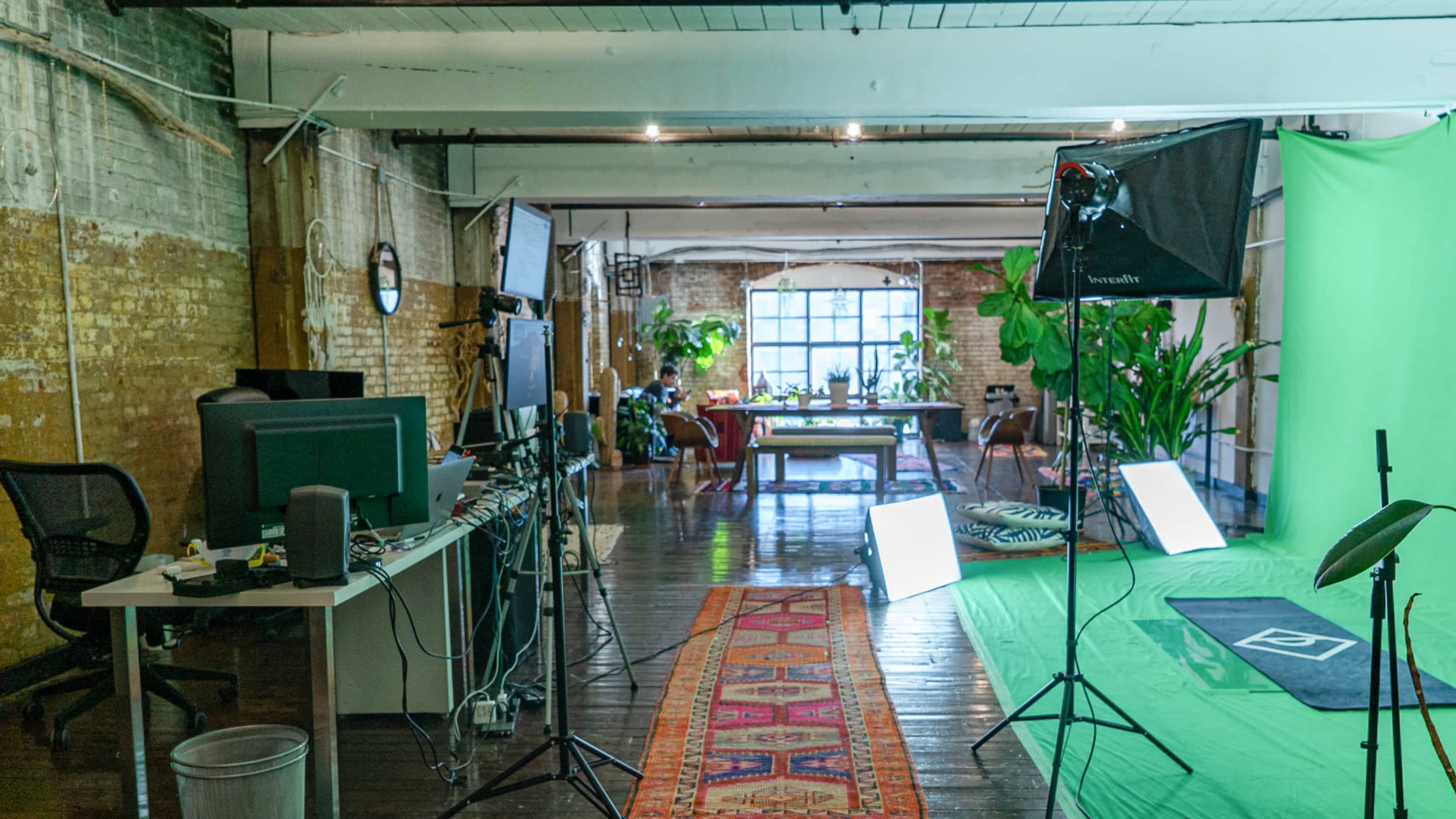 The image shows a bright studio space with a green screen setup, various plant decorations, and multiple workstations arranged in an open layout.