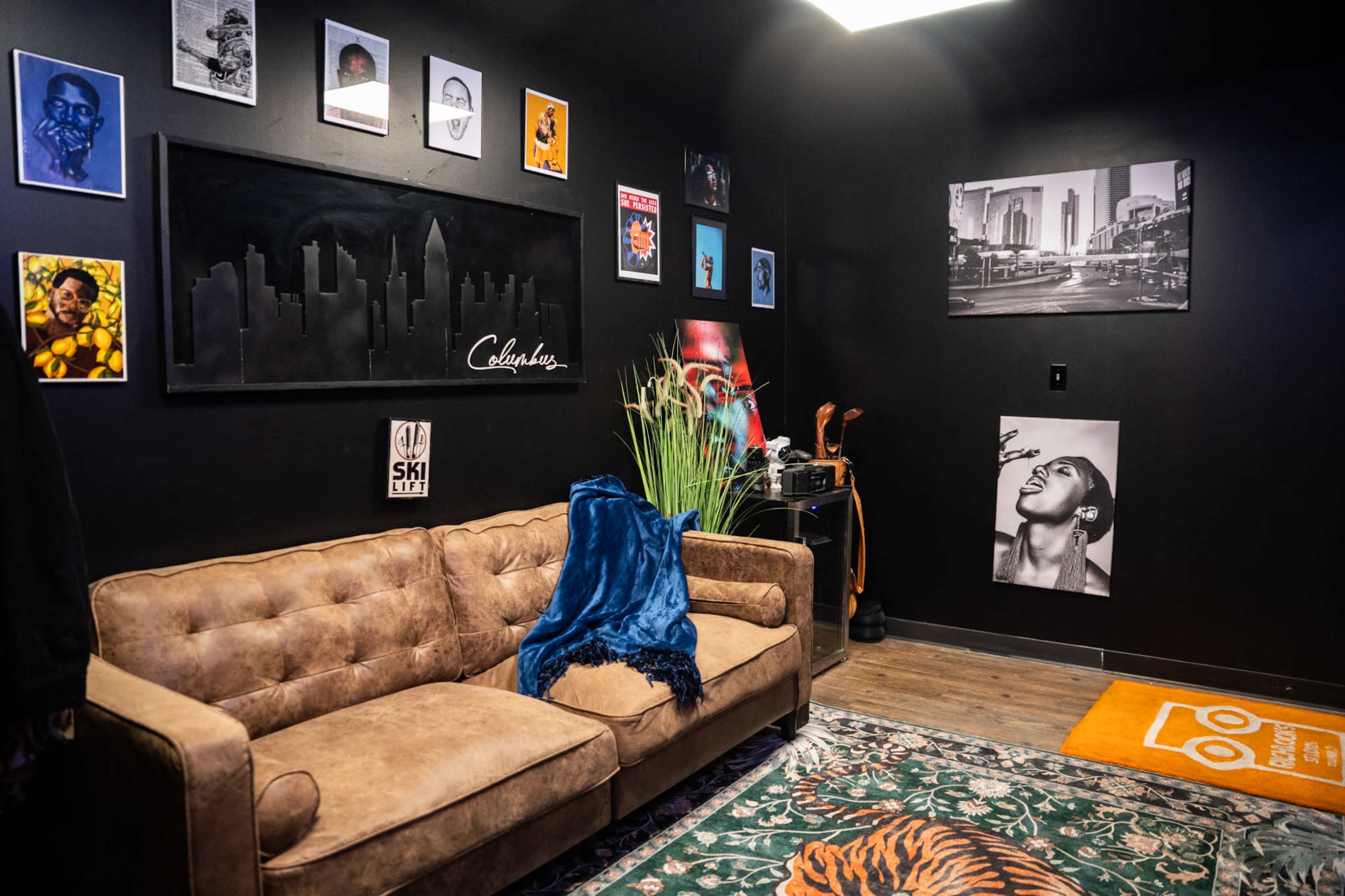The image shows a dimly lit room featuring a brown tufted sofa, a patterned rug with a tiger design, and various framed artworks hanging on the black walls.