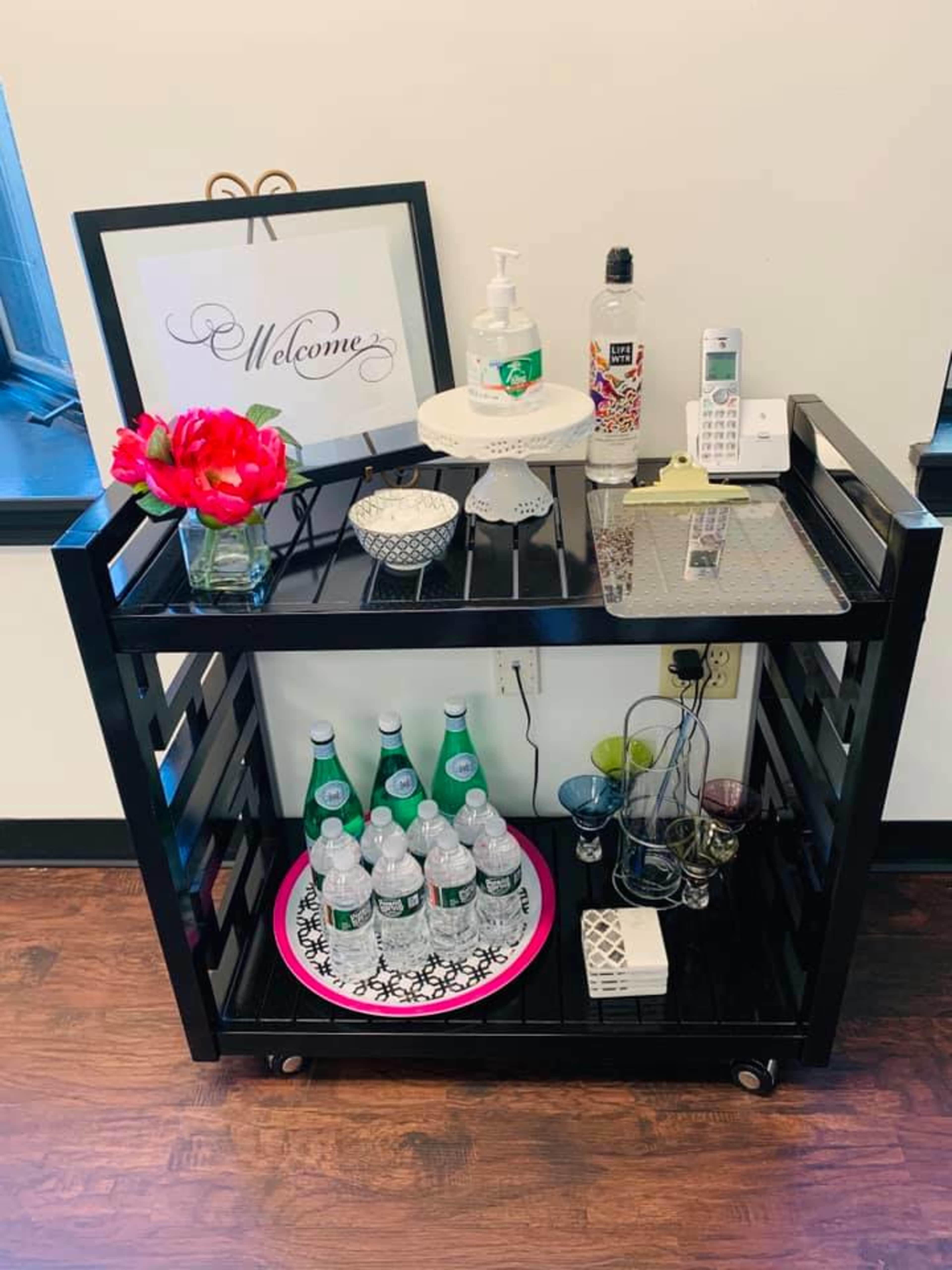 A black cart is set up with decorative items, including flowers, beverages, and hand sanitizer, in a well-lit space.