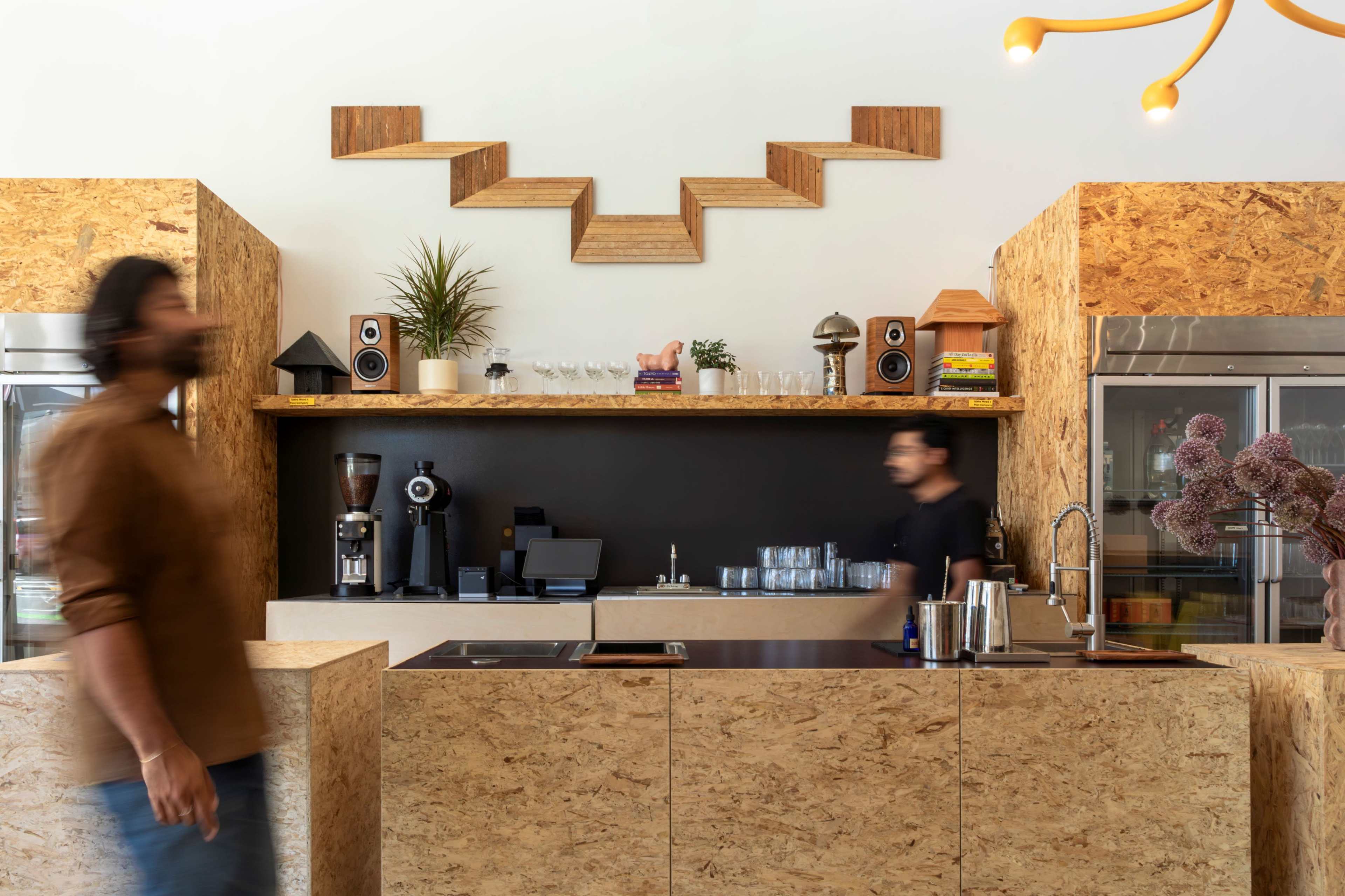 The image shows a modern coffee shop interior featuring a wooden counter, decorative wall art, and two staff members engaged in their tasks.