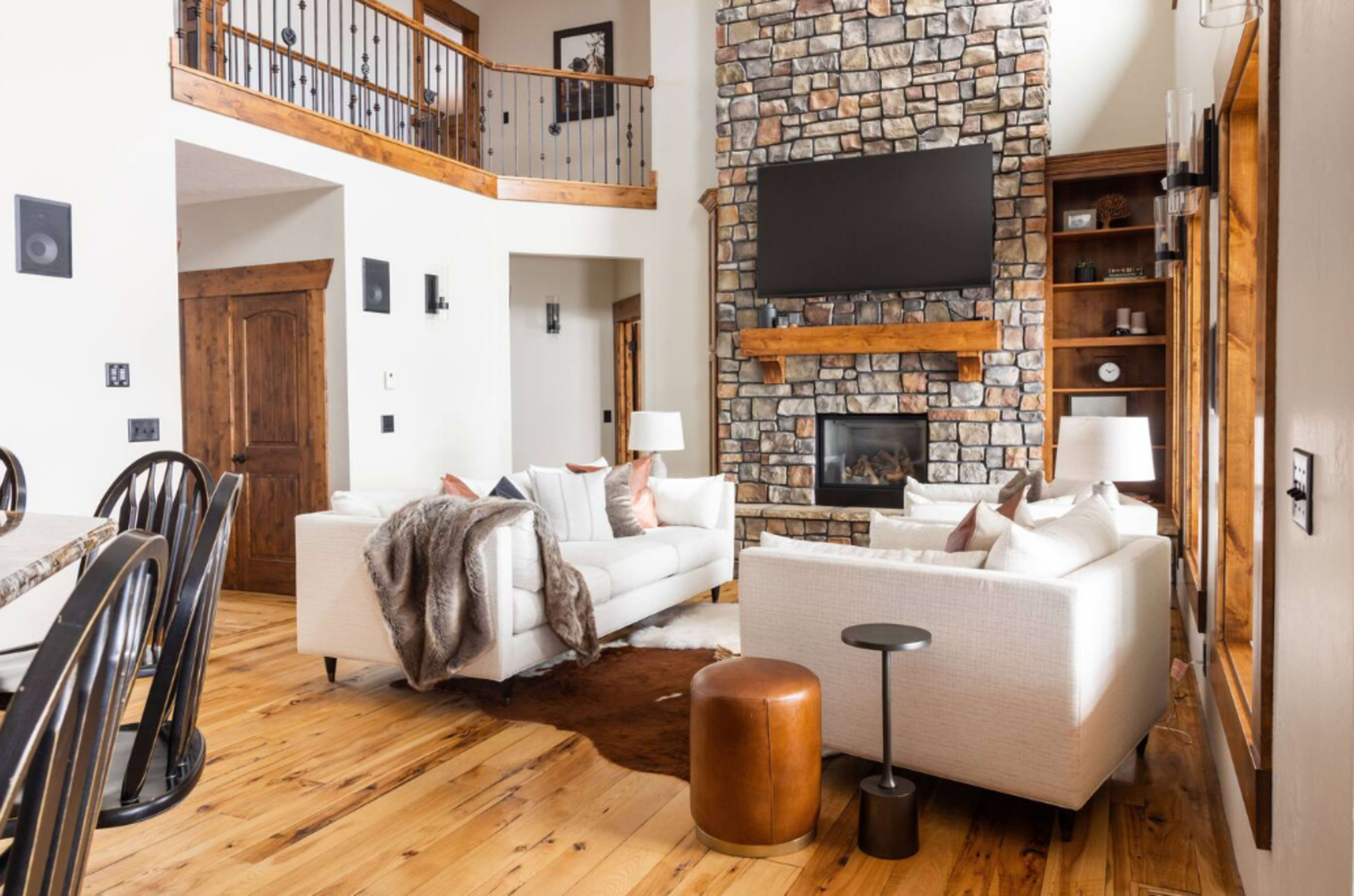 The image shows a modern living room with a stone fireplace, a sectional sofa, and wooden accents, along with a dining area featuring a large table.