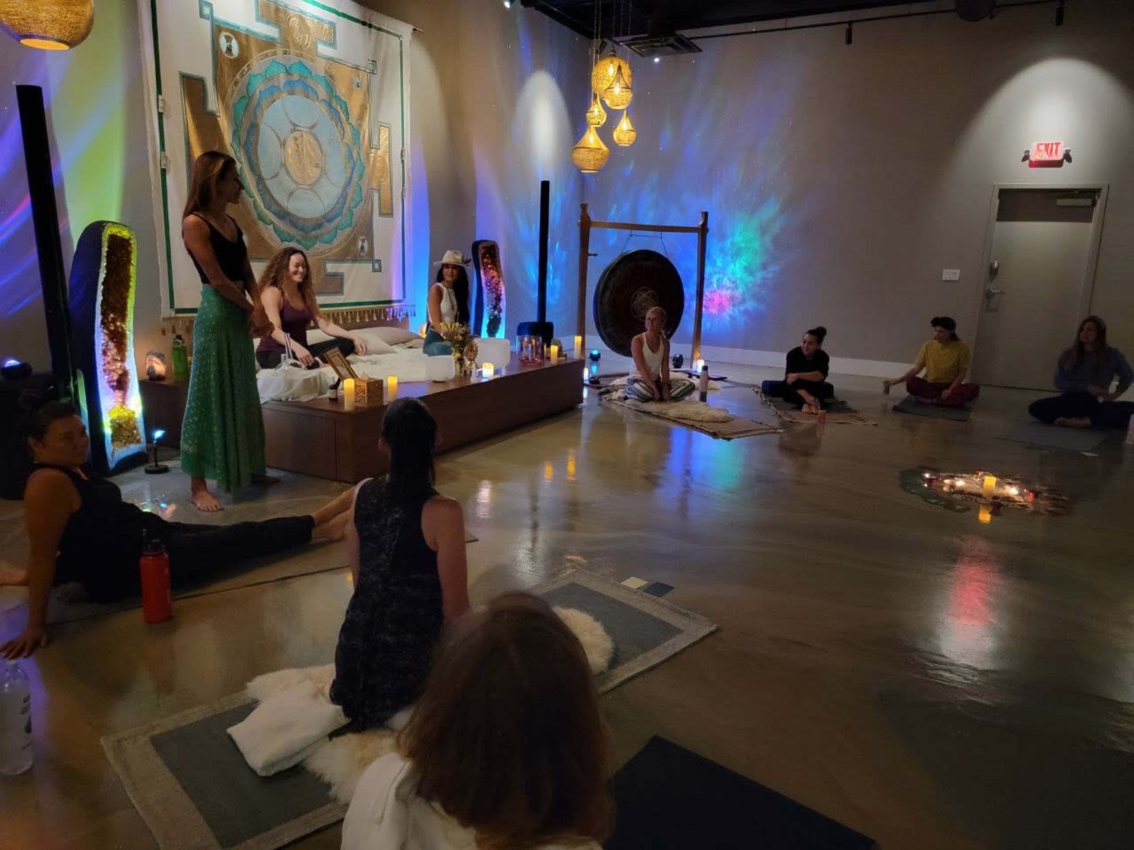 A group of people sit on the floor in a dimly lit room arranged for a wellness or meditation session, with colorful lights and decorative elements in the background.