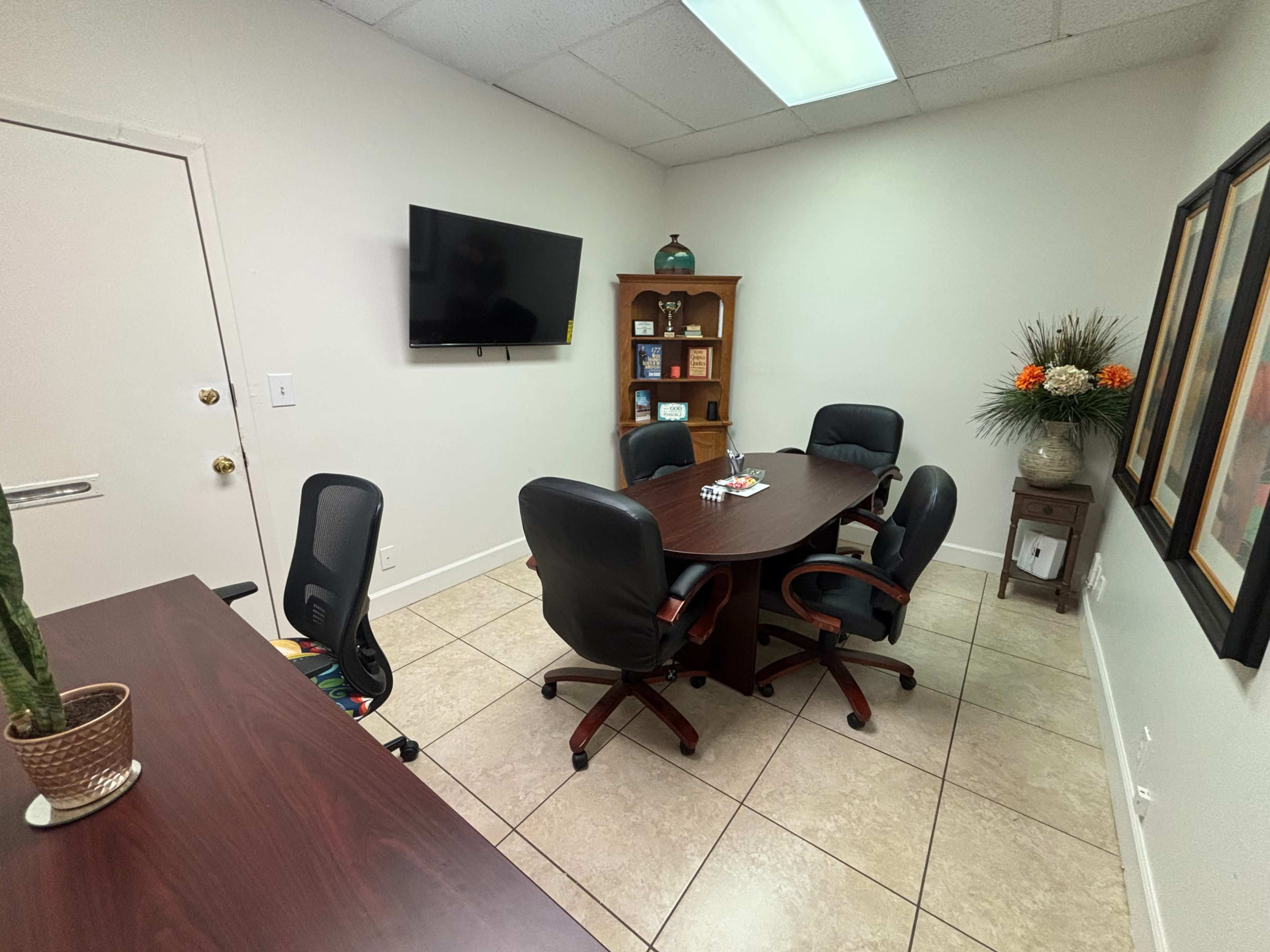 The image shows a small conference room with a circular table surrounded by four chairs, a wall-mounted TV, and decor including a plant and framed artwork.